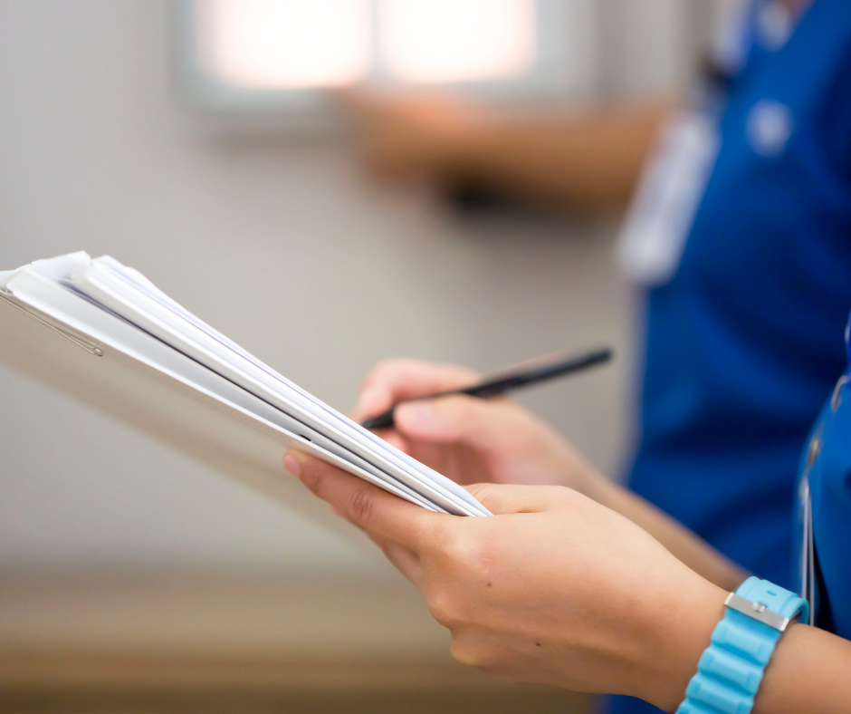 Person in scrubs holding a clipboard and pen, writing notes.