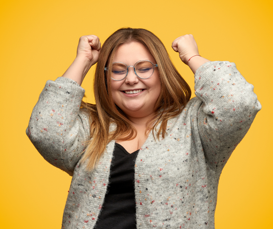 Woman smiling with arms raised against yellow background, symbolizing the confidence and joy of living agefully through Second Act Women