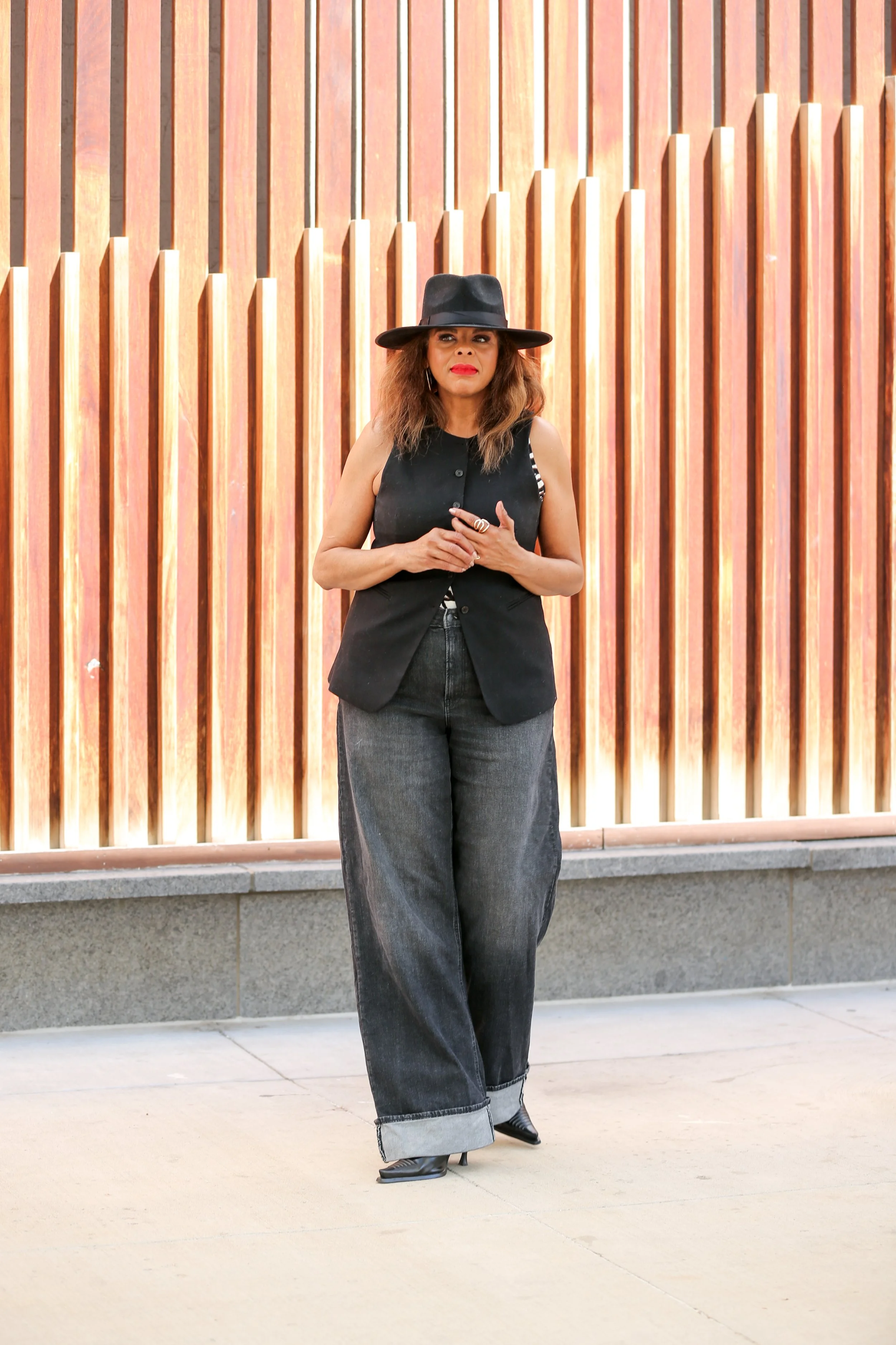 Barbara Brooks, founder of Second Act Women standing outdoors in front of a wooden slat wall, wearing a black sleeveless vest, large black hat, wide-leg jeans with cuffs, and black boots -- standing confidently reflecting Second Act Women.