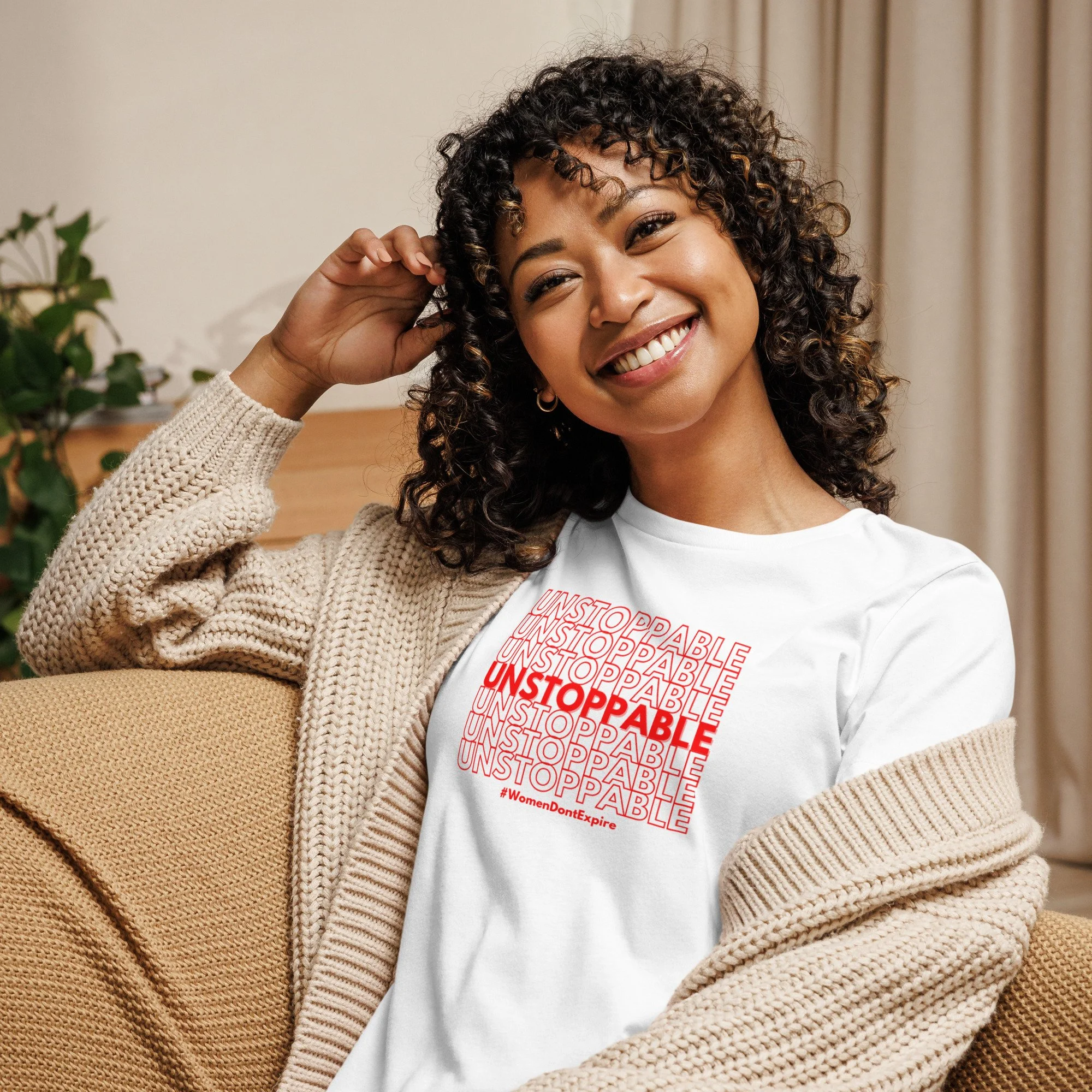 A smiling woman with curly hair wearing a white t-shirt with the word "UNSTOPPABLE" in red, a beige knitted cardigan, and hoop earrings, sitting on a tan sofa near a plant and curtains.