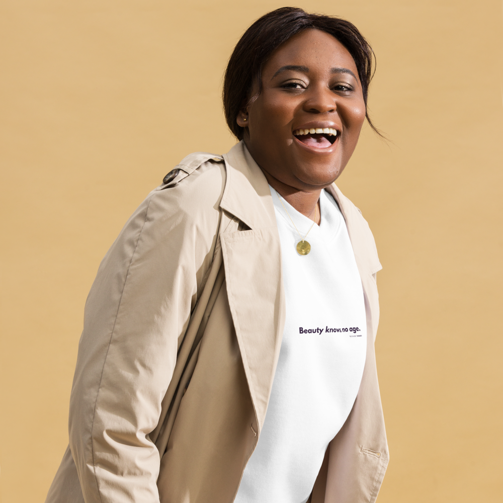 Woman with short dark hair smiling in a beige trench coat and a white tee that reads Beauty knows no age. Represents the Barbara Brooks and SecondActWomen message of living agefully.