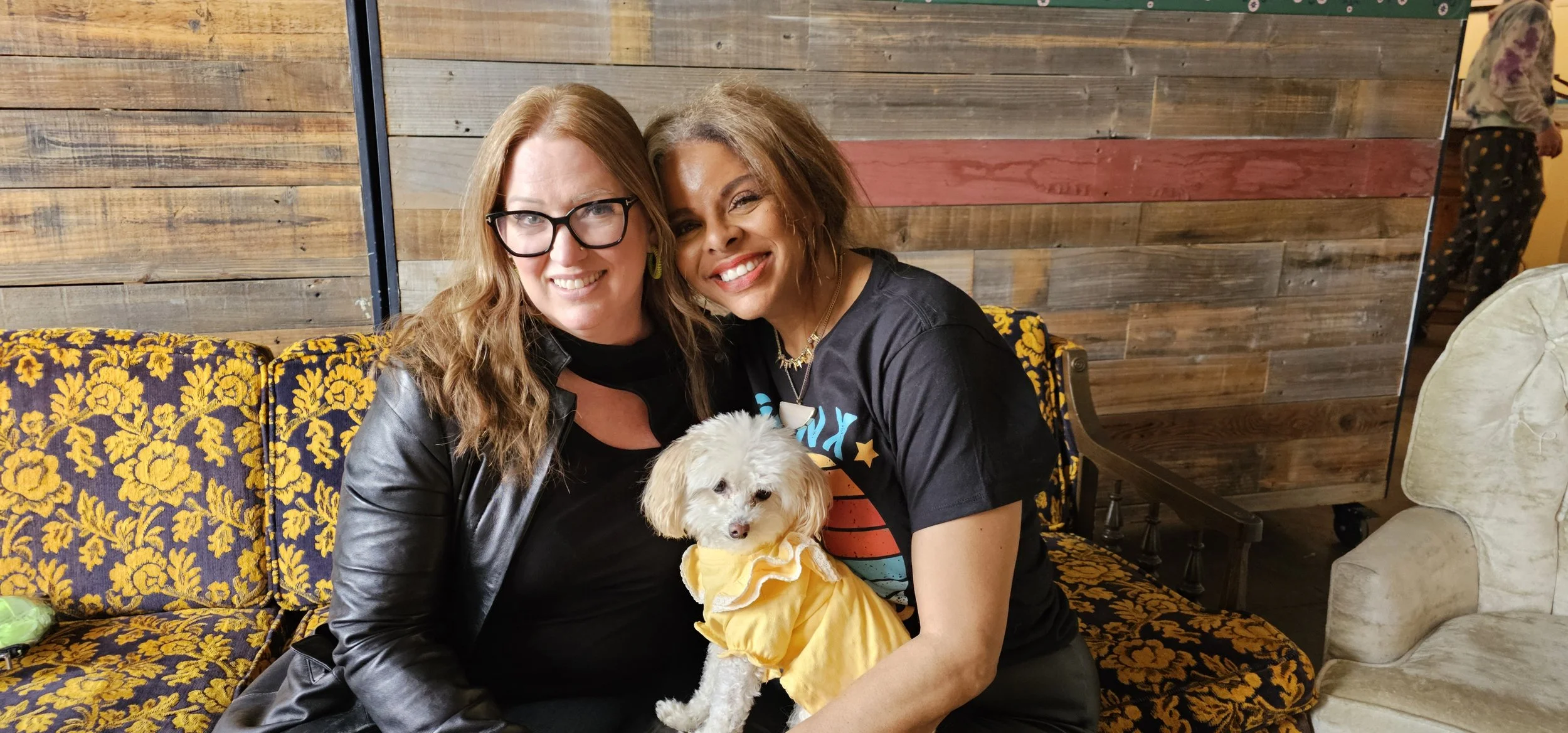 Two women sitting on a floral-patterned couch, smiling, with a small white dog wearing a yellow dress, in a wooden interior setting.