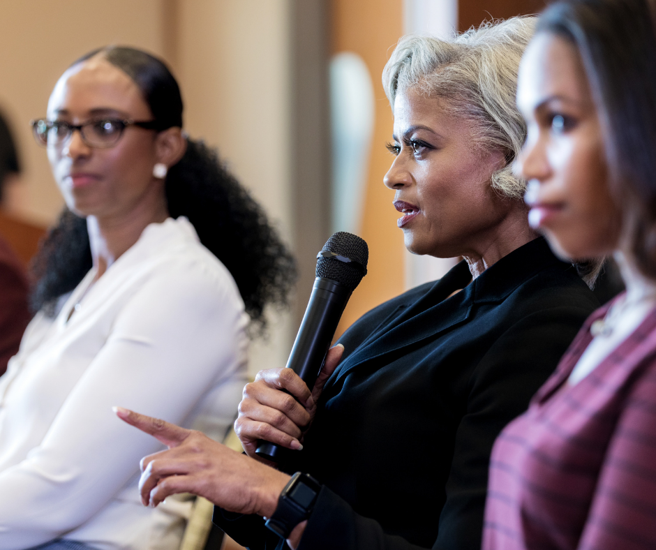 Women attending a panel discussion, one woman speaking into a microphone and others listening.