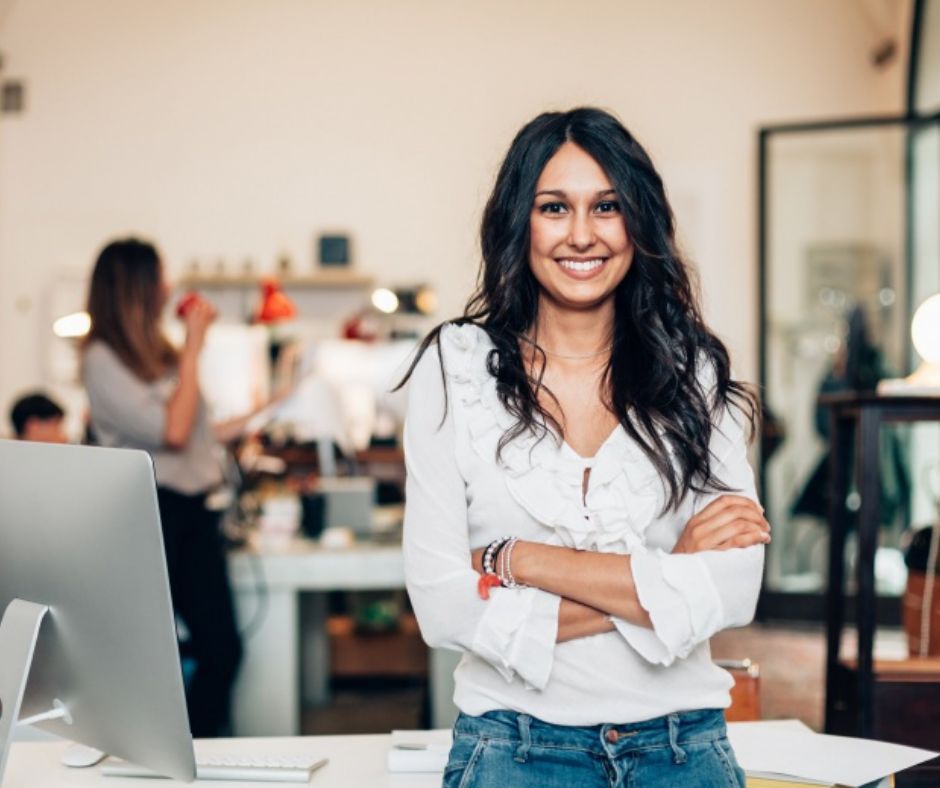 Smiling woman with dark wavy hair standing in an office or creative workspace with others in the background.