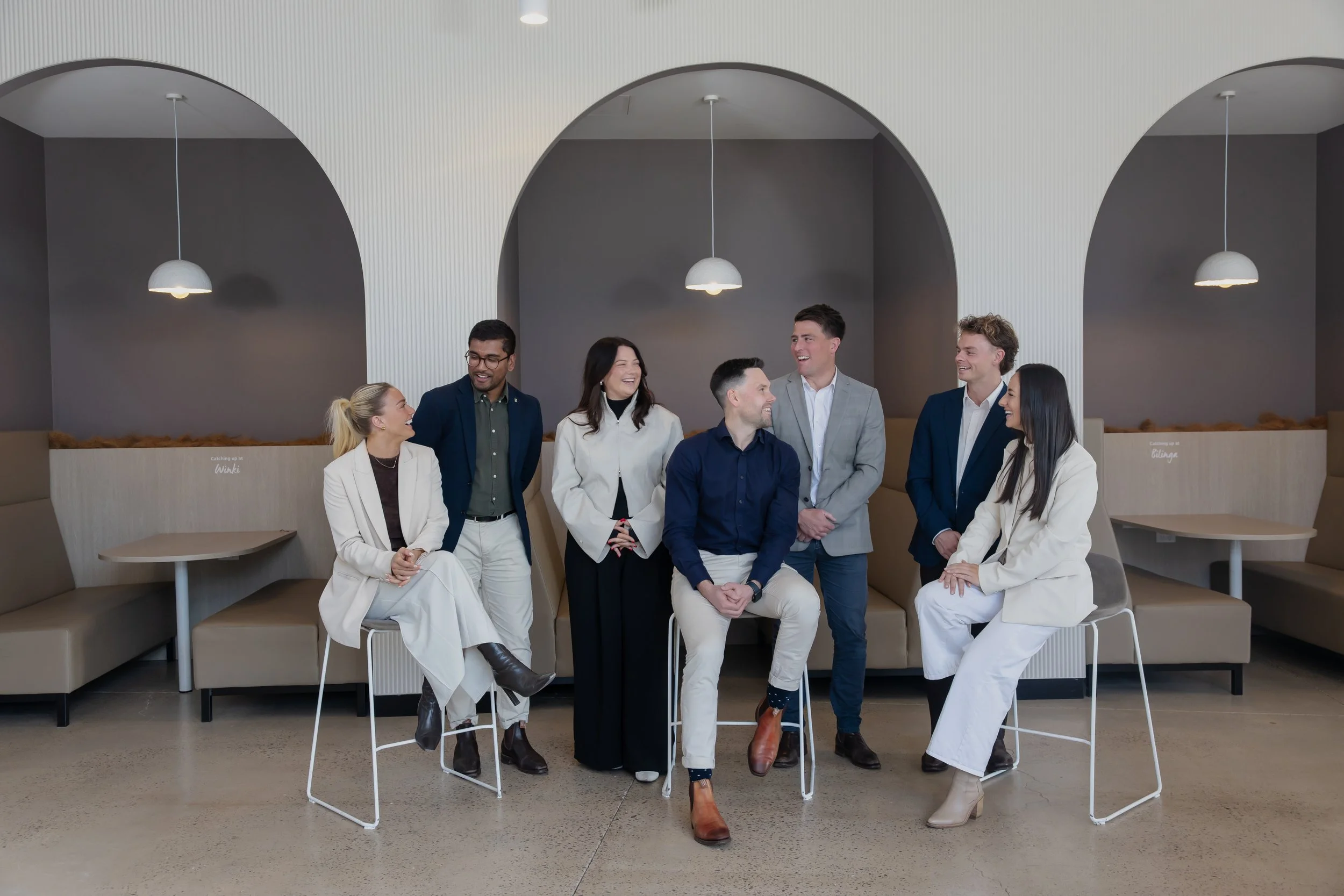 A group of seven diverse young adults, four women and three men, gathered in a modern, stylish restaurant or café with arched ceilings and hanging pendant lights. They are engaged in conversation, smiling and laughing, with some seated and others sta