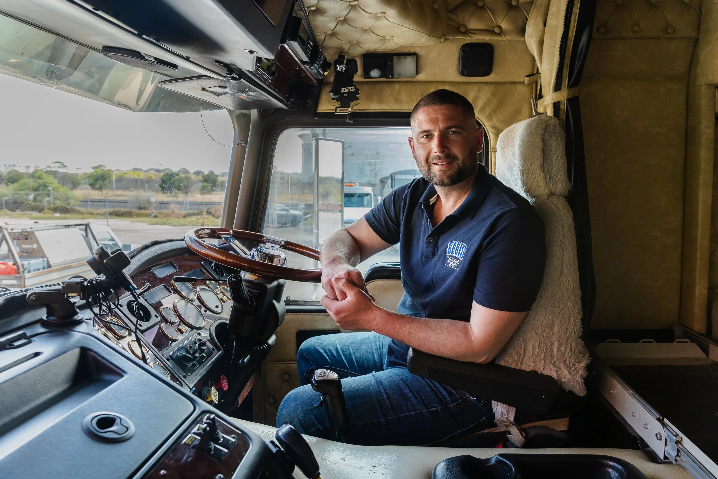 A man with short hair and a beard sitting in the driver's seat of a large truck, smiling and looking at the camera, with a view of the outside highway through the windshield.
