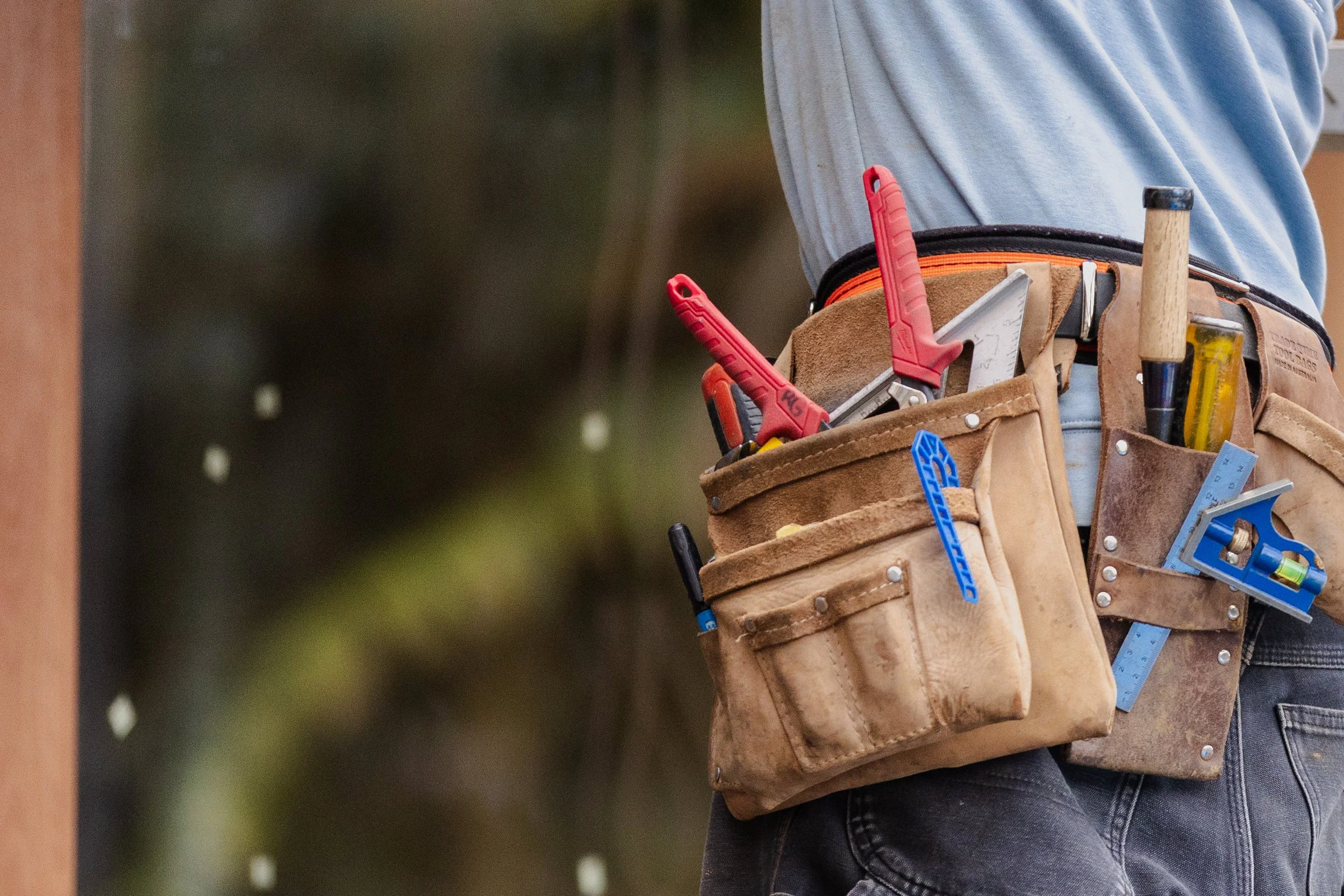 Construction worker's brown leather tool belt containing various tools such as pliers, a hammer, a level, and a measuring tape, with a person wearing a gray shirt and jeans.