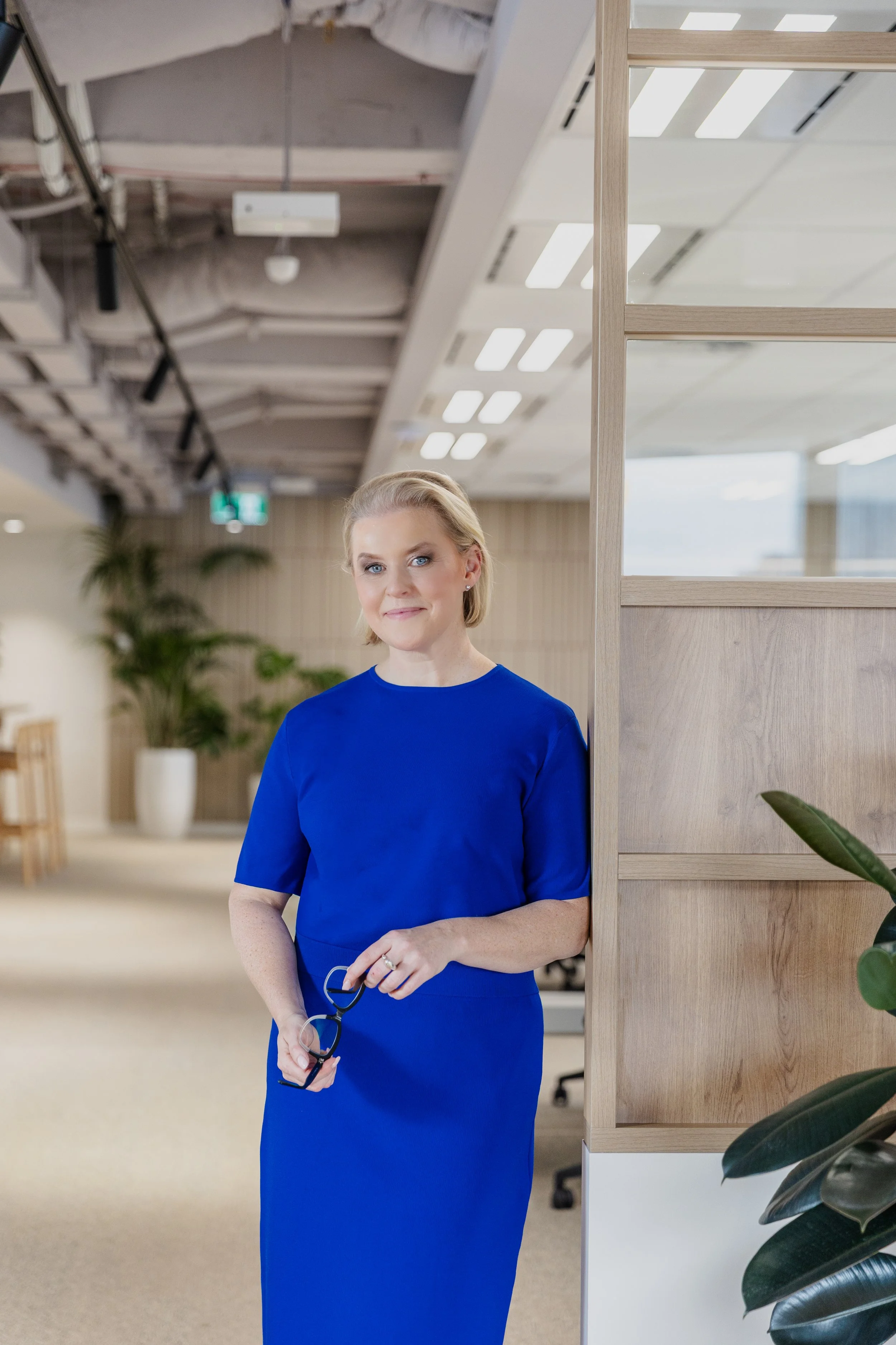 A woman with blonde hair wearing a royal blue dress, standing in an office environment, holding glasses and smiling.