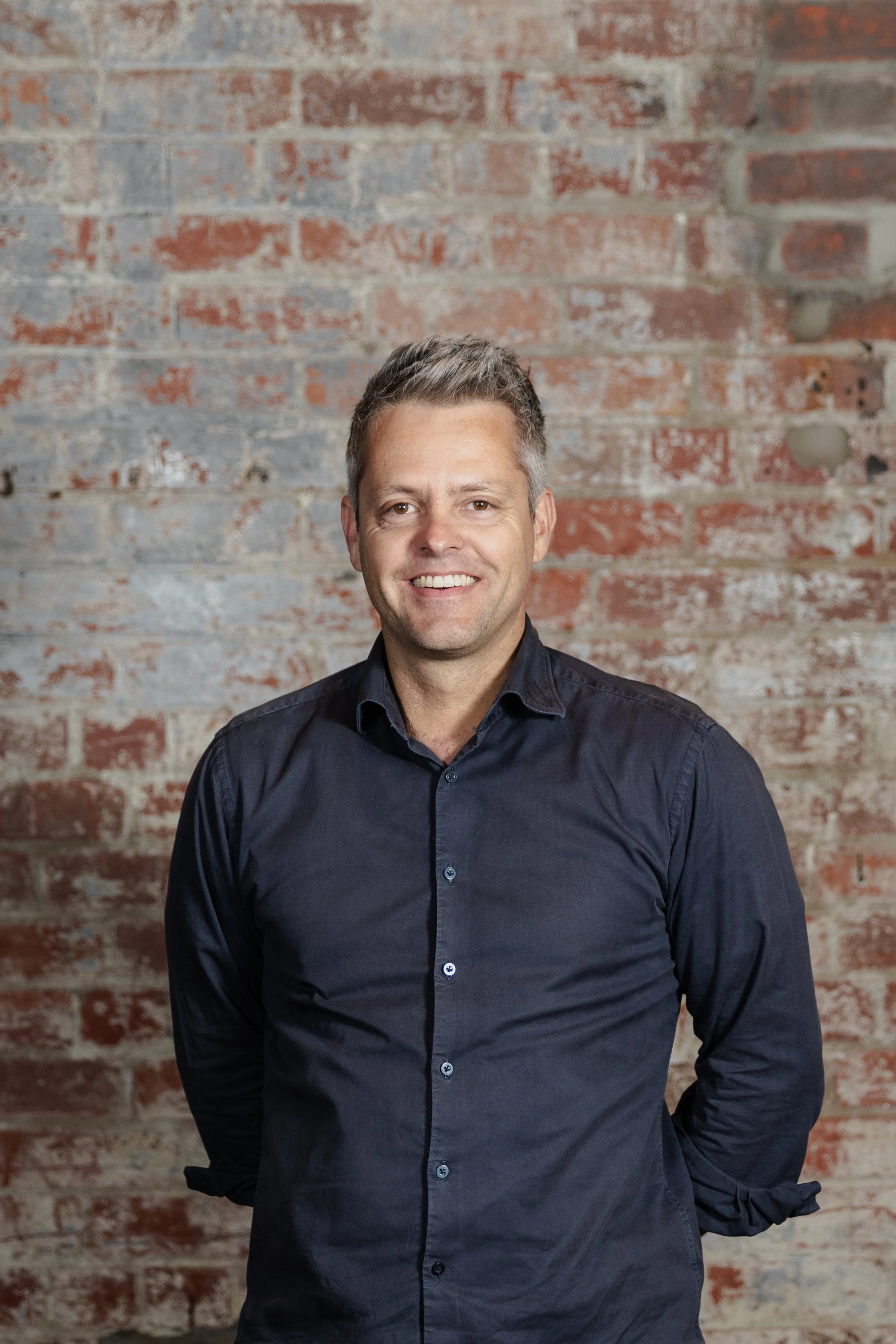 A man smiling in front of a brick wall, wearing a dark button-up shirt.