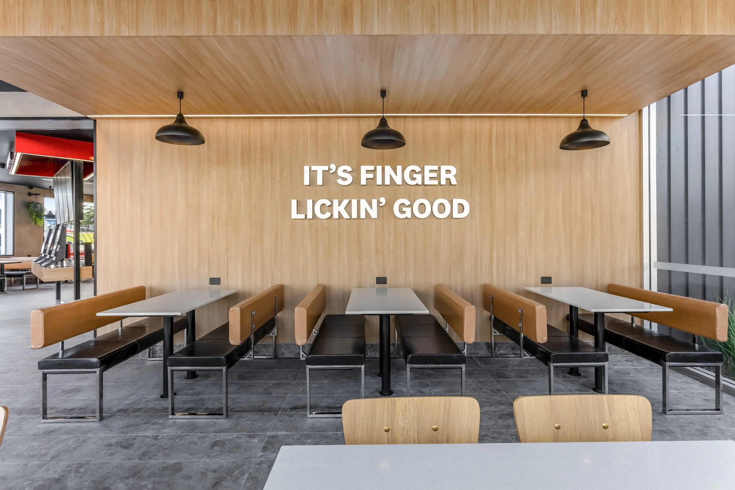  Interior of a restaurant with wood-paneled walls, black pendant lighting, and empty tables with black and wooden chairs. There is a sign on the wall that says, "It’s finger lickin’ good."