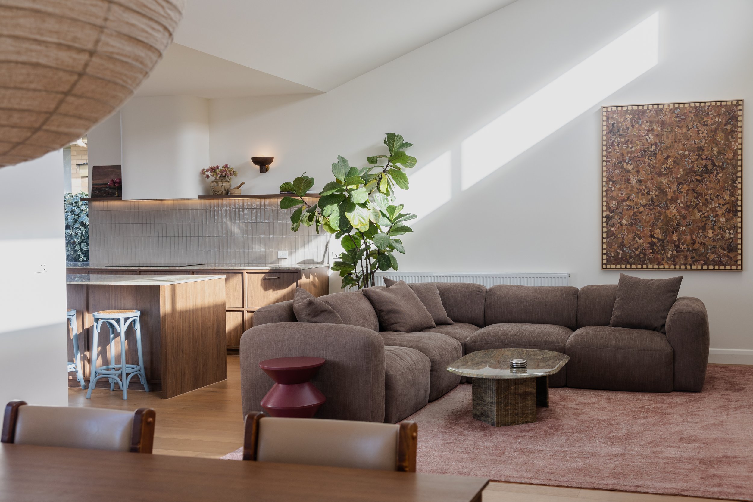 A cozy living room with a sectional sofa and a small marble-top coffee table, a plant, and an abstract wall hanging, illuminated by natural sunlight.
