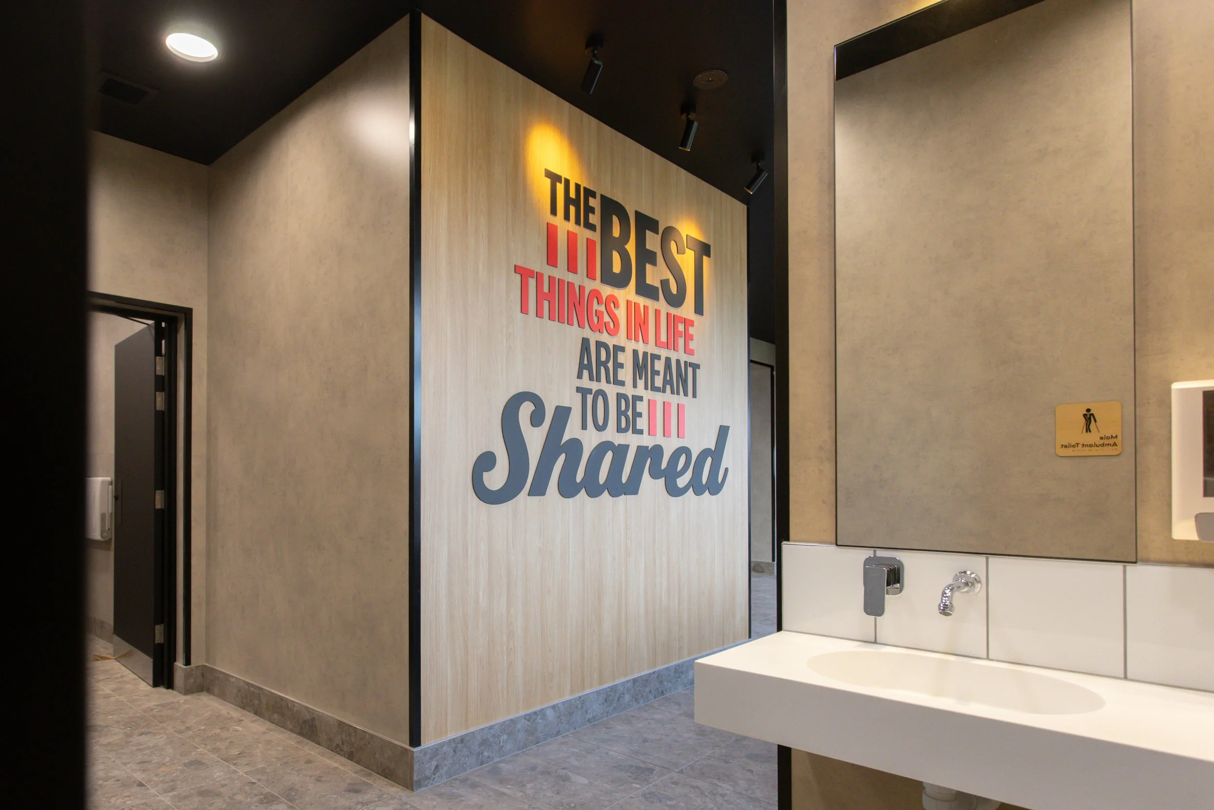 Bathroom with a wooden wall featuring a large sign that reads 'The Best Things in Life Are Meant to Be Shared' in colorful letters; mirror and sink visible on right side; door and partial wall on left side.