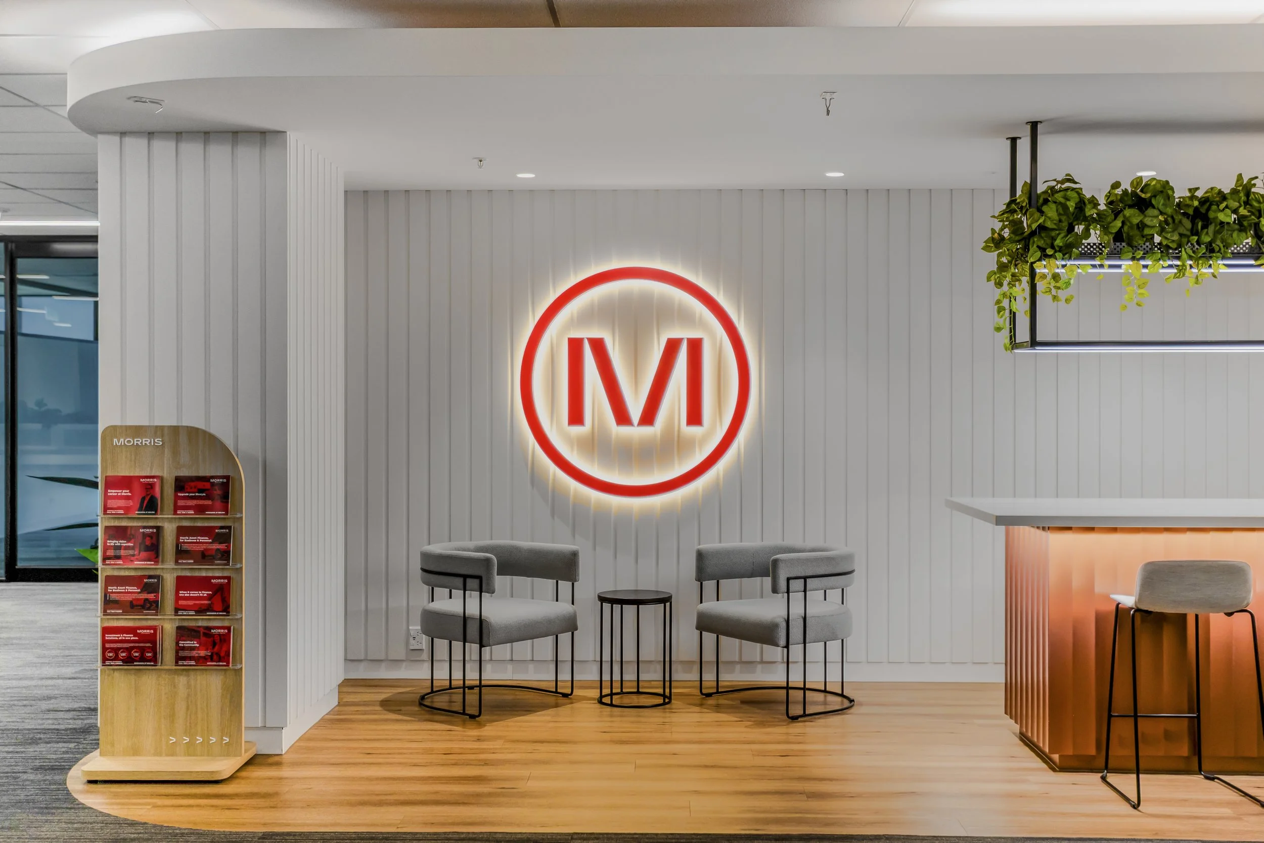 Modern interior space with a red outlined letter 'M' lit sign on a white paneled wall, surrounded by seating and greenery.