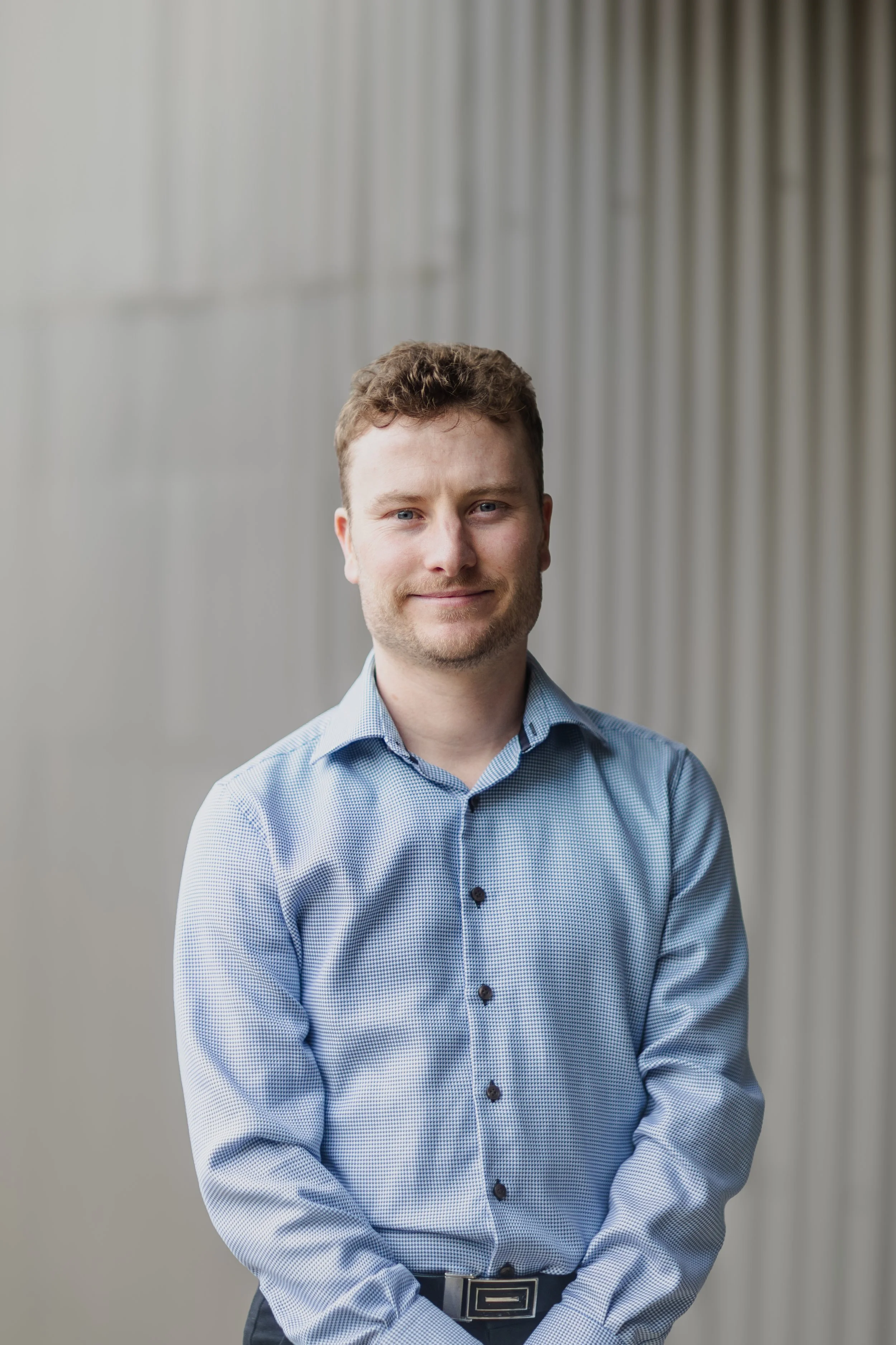 A young man with short, curly brown hair and blue eyes stands in front of a neutral background with vertical lines. He is smiling slightly and wearing a light blue dress shirt with dark buttons and a black belt.