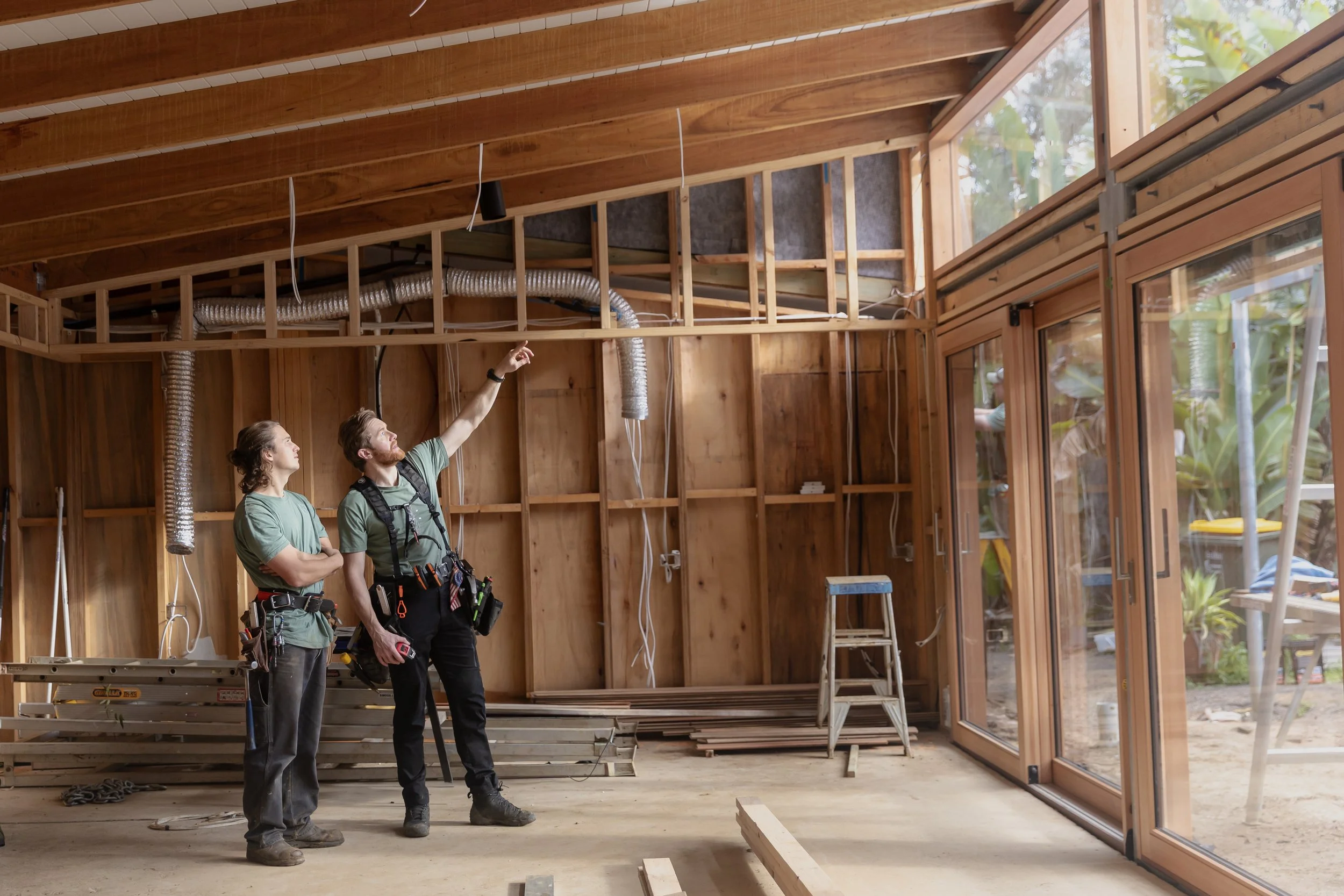 Two workers inside a construction site, looking at the ceiling with wooden beams, ventilation duct, and large windows.