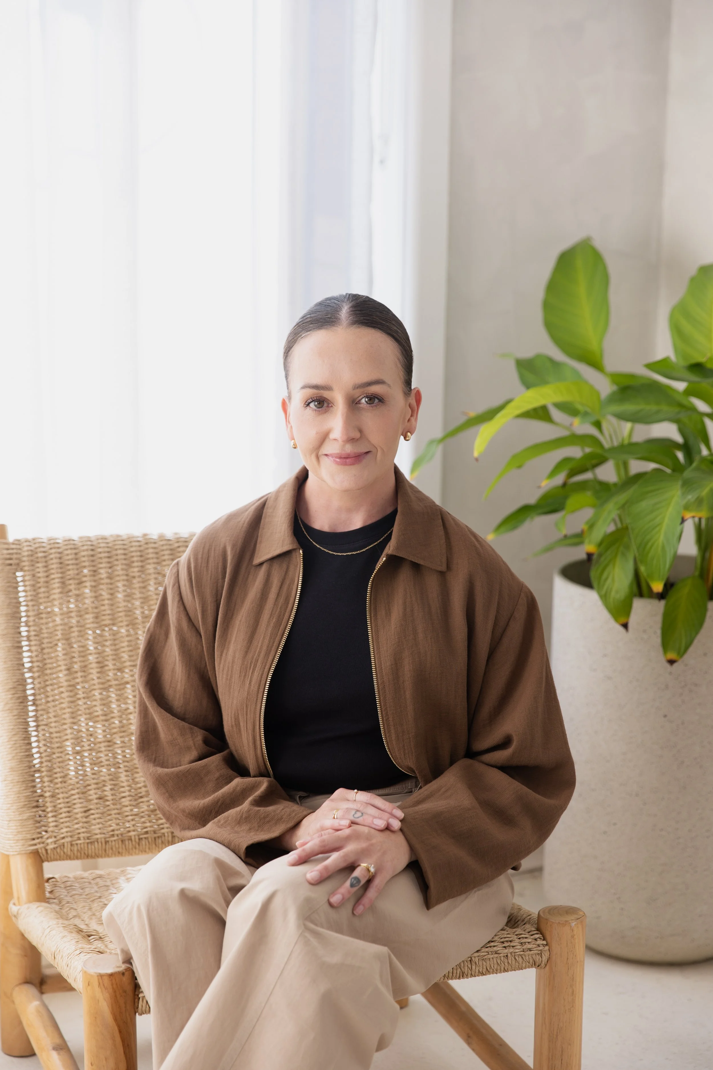 A woman with dark hair pulled back, sitting on a woven chair in front of a light background with sheer curtains and a large green houseplant, wearing a black top and a brown jacket.