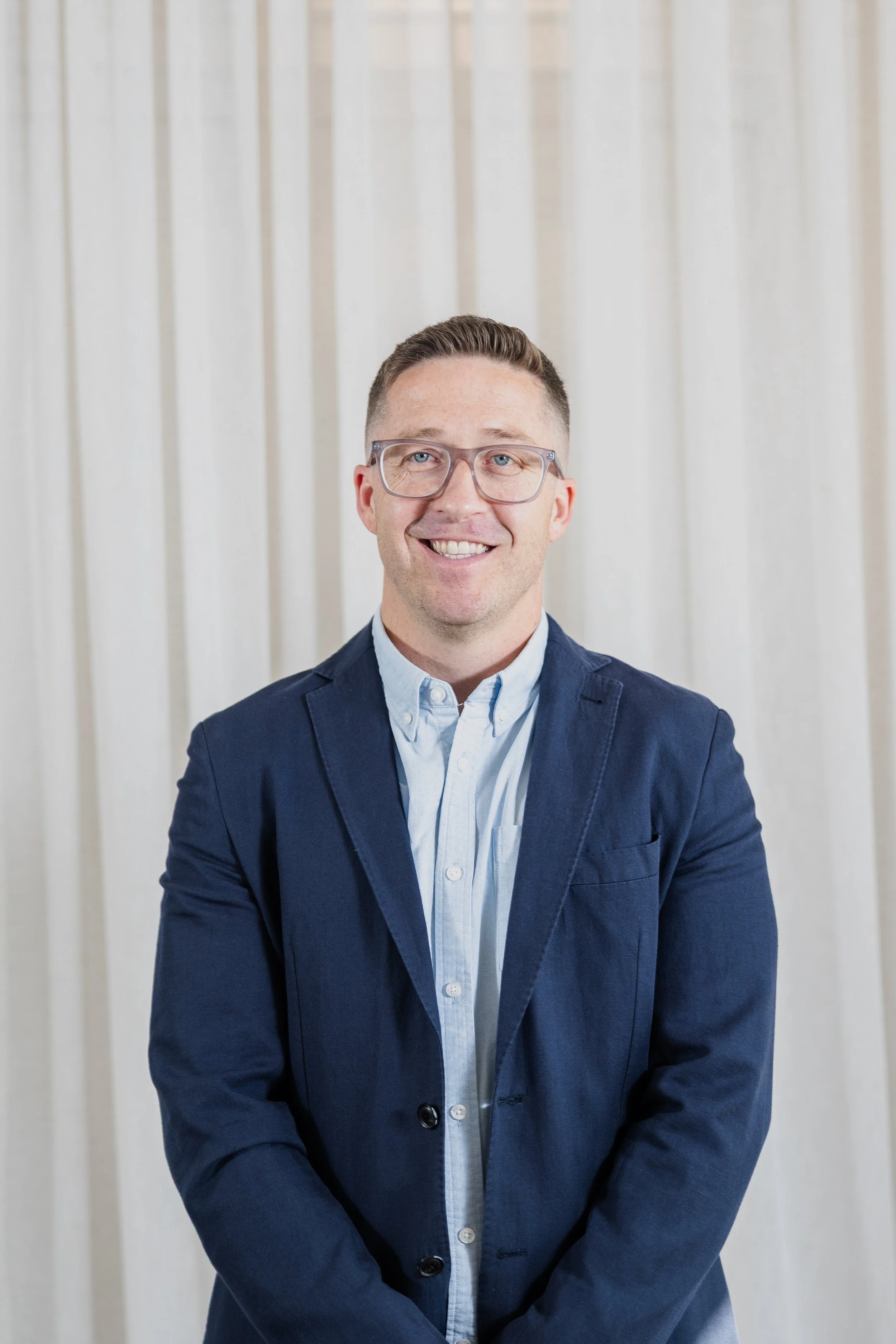 A smiling man with glasses wearing a navy blue blazer and light blue shirt. He is standing in front of a neutral-colored curtain background.