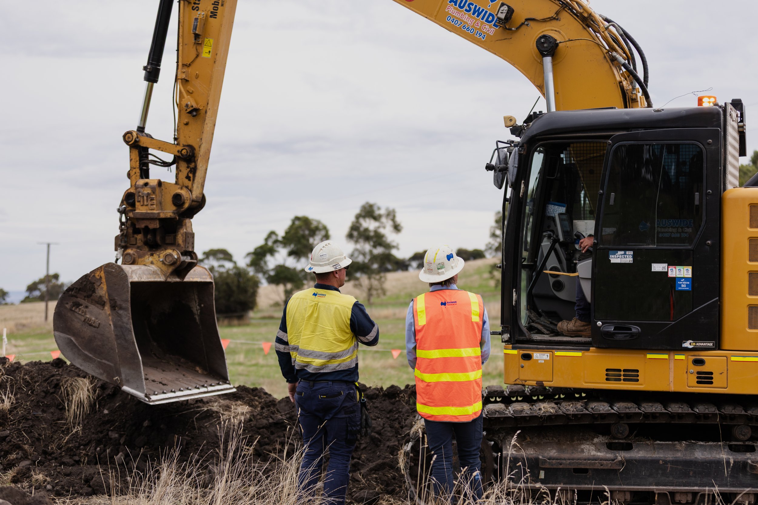 Construction workers in hard hats and safety vests observe a yellow excavator digging into the ground at a construction site under a cloudy sky.