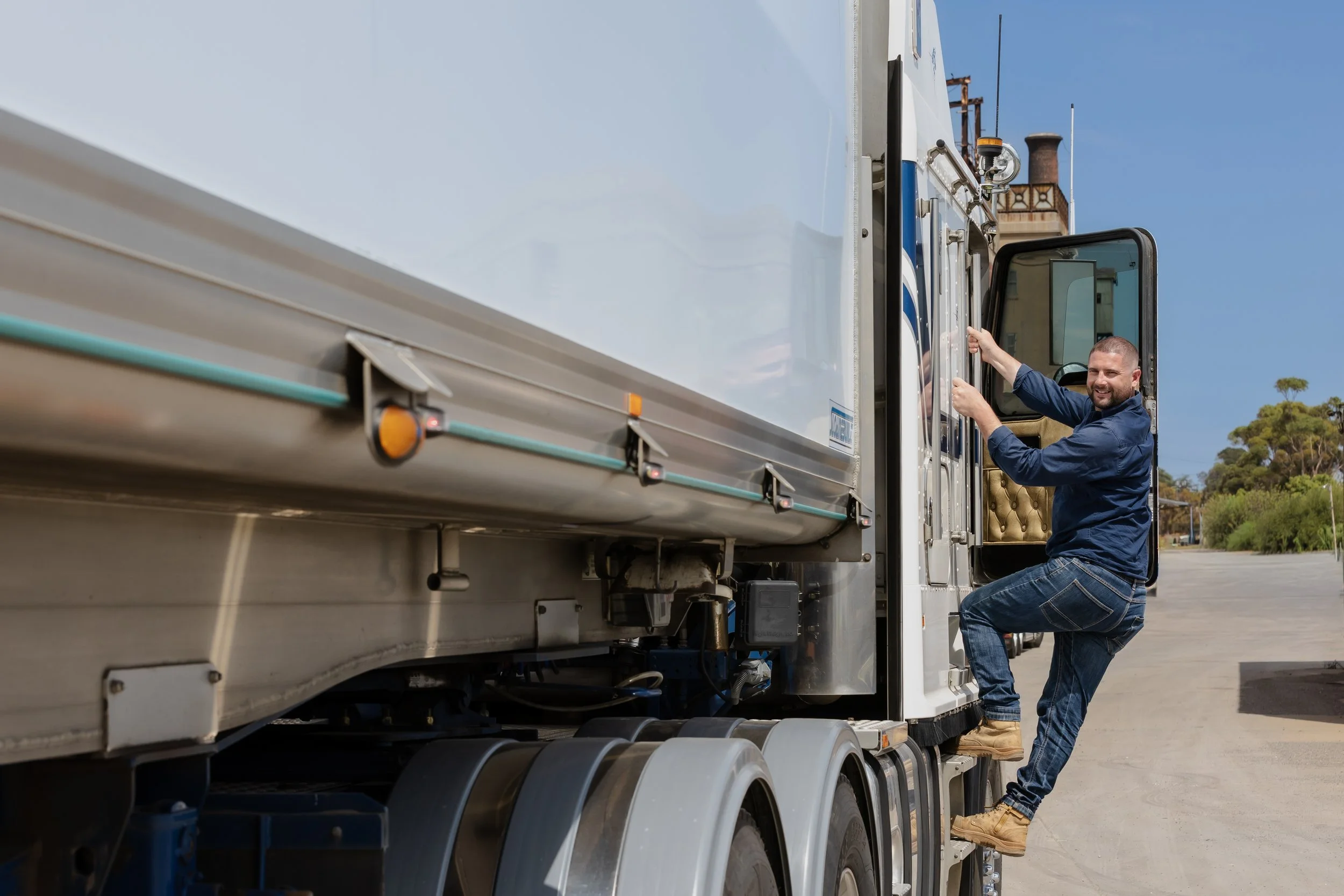 A man with a beard, wearing a blue shirt and jeans, is climbing into the cab of a large white semi-truck or tractor-trailer. The truck is parked outdoors under a clear blue sky, with some trees and a smoke stack visible in the background.