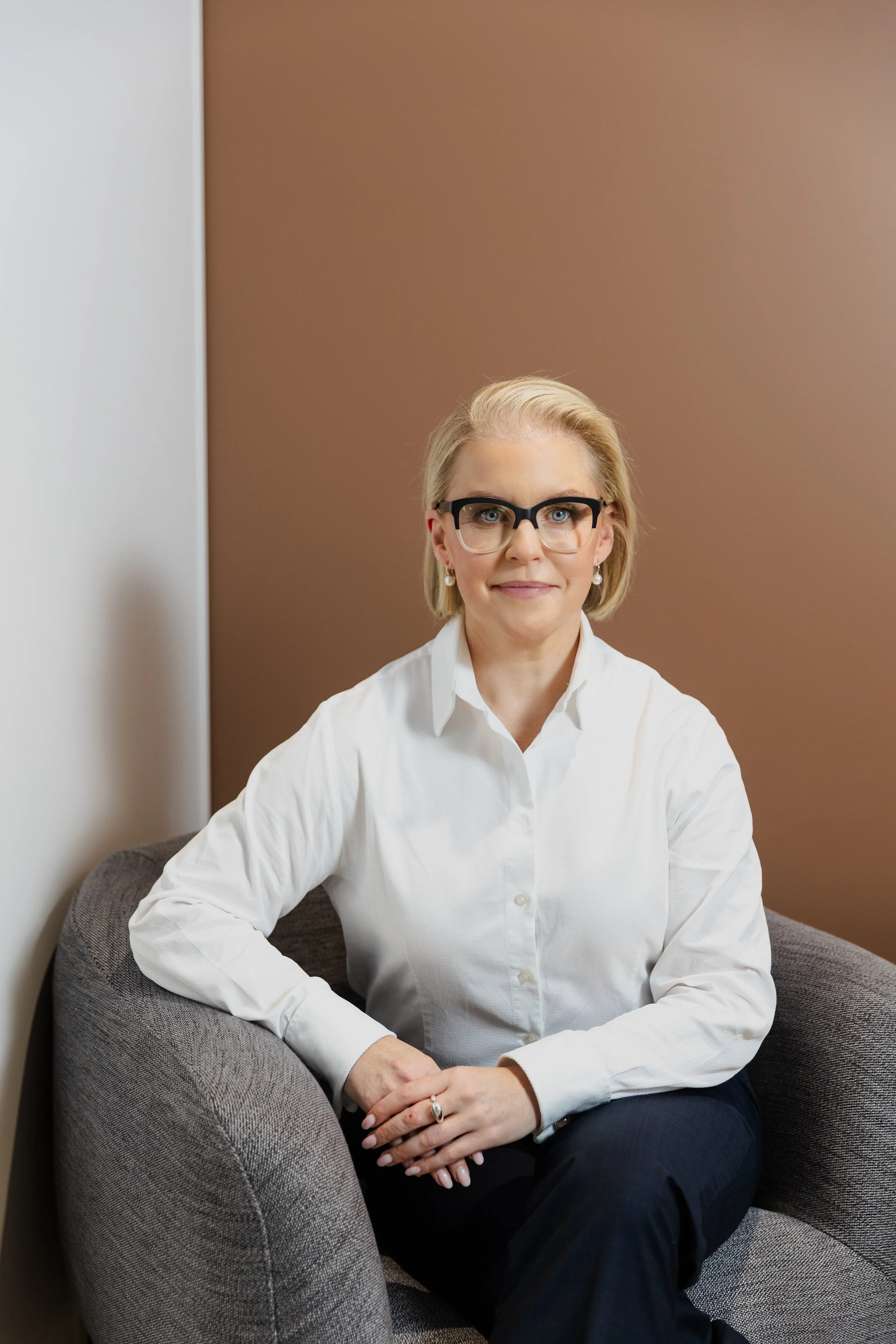 A woman with blonde hair, glasses, and pearl earrings sitting on a gray upholstered chair against a brown and white wall, wearing a white blouse.