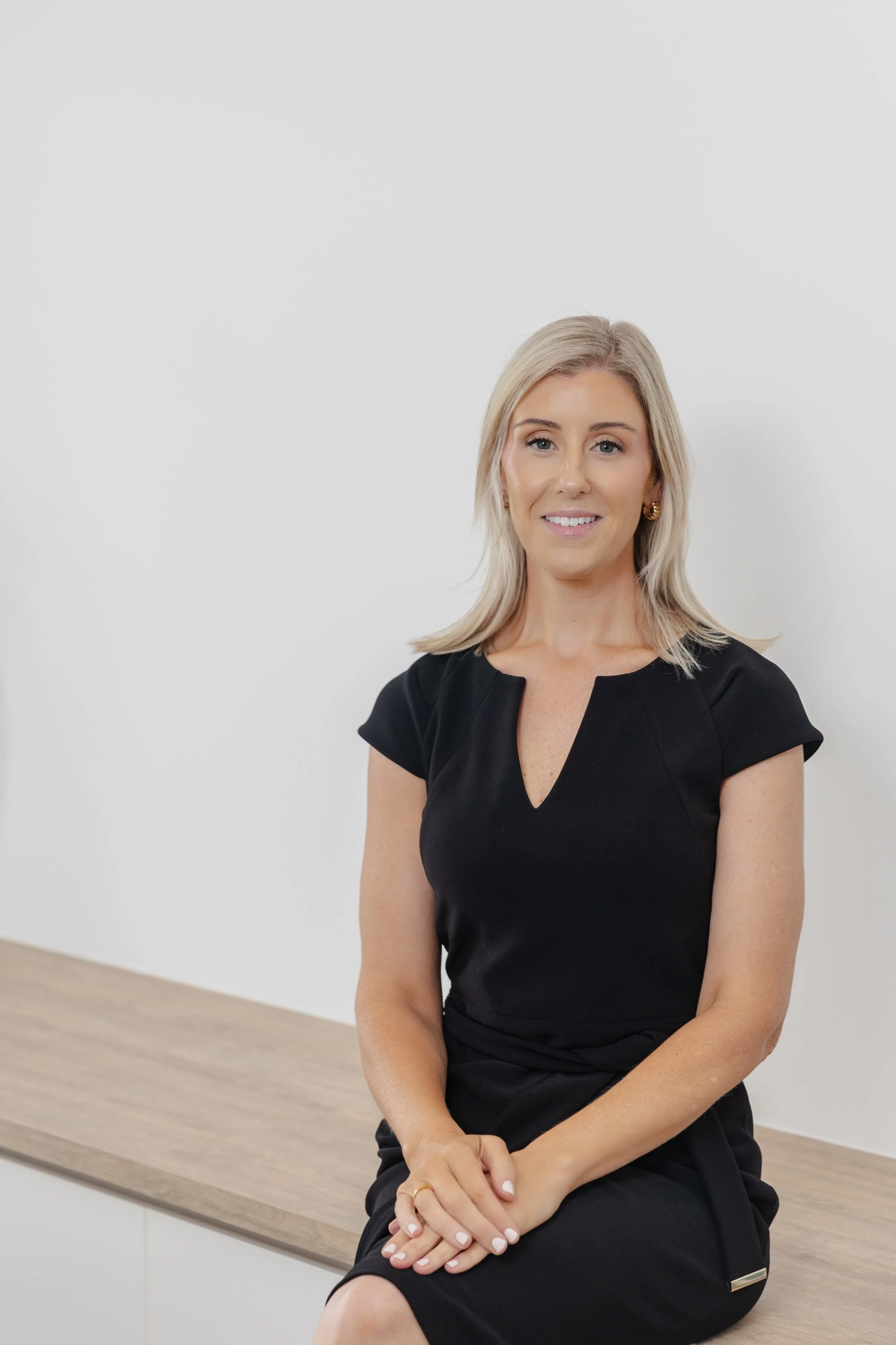 A woman with blonde hair, wearing a black dress, sitting on a wooden bench against a white wall, smiling at the camera.