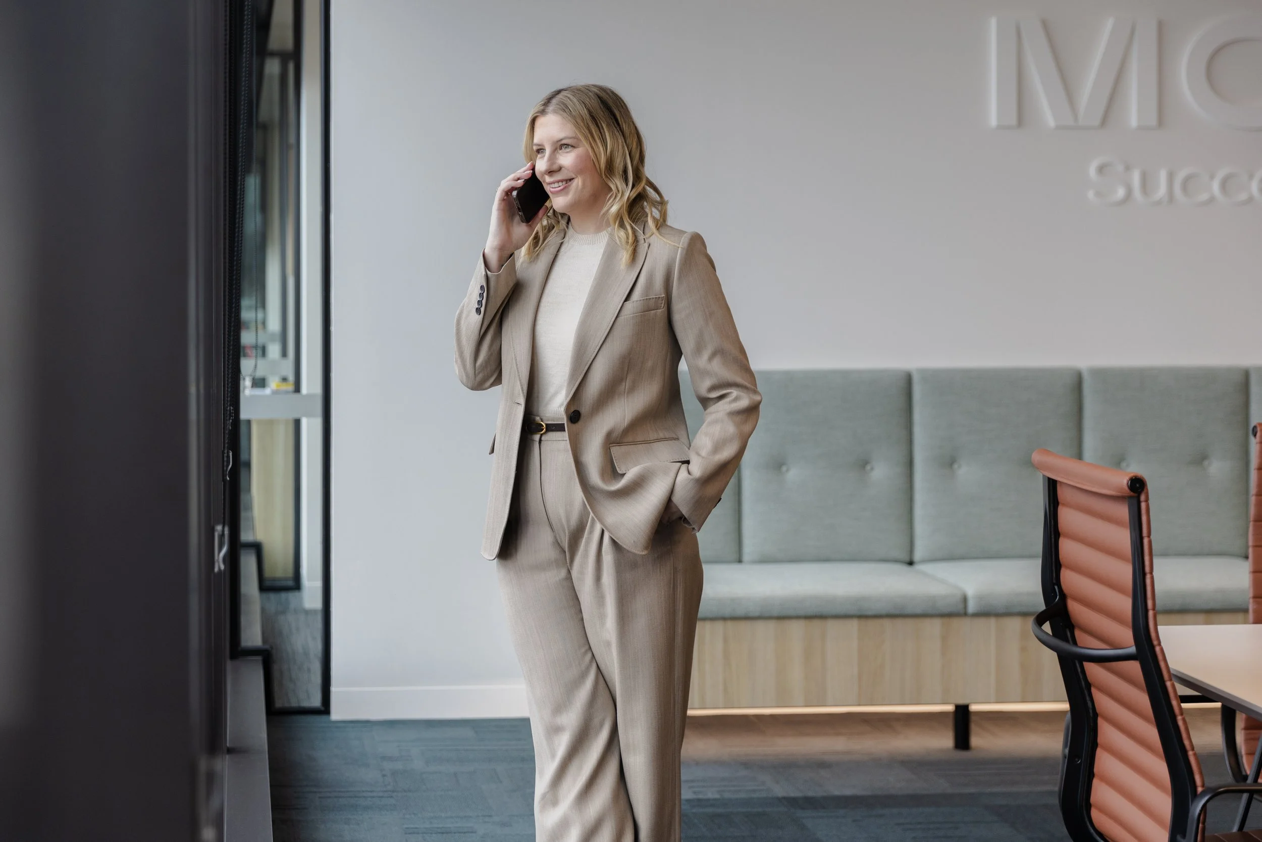 A woman with blonde hair wearing a beige business suit talking on her cell phone in an office lobby area.