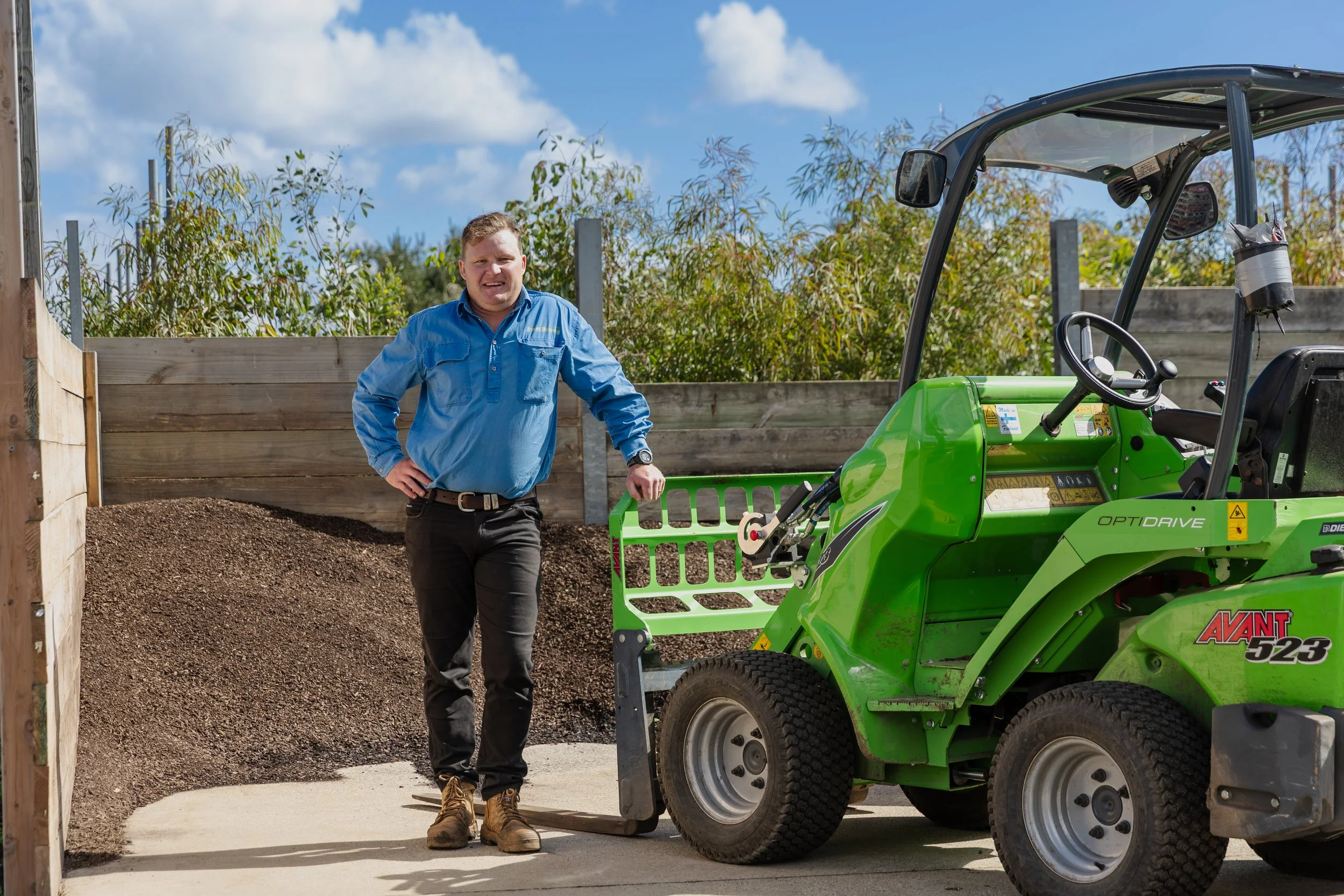 A man in a blue shirt and black pants stands beside a green compact utility tractor with a front loader attachment on a construction site. There is a wooden fence and trees in the background under a partly cloudy sky.