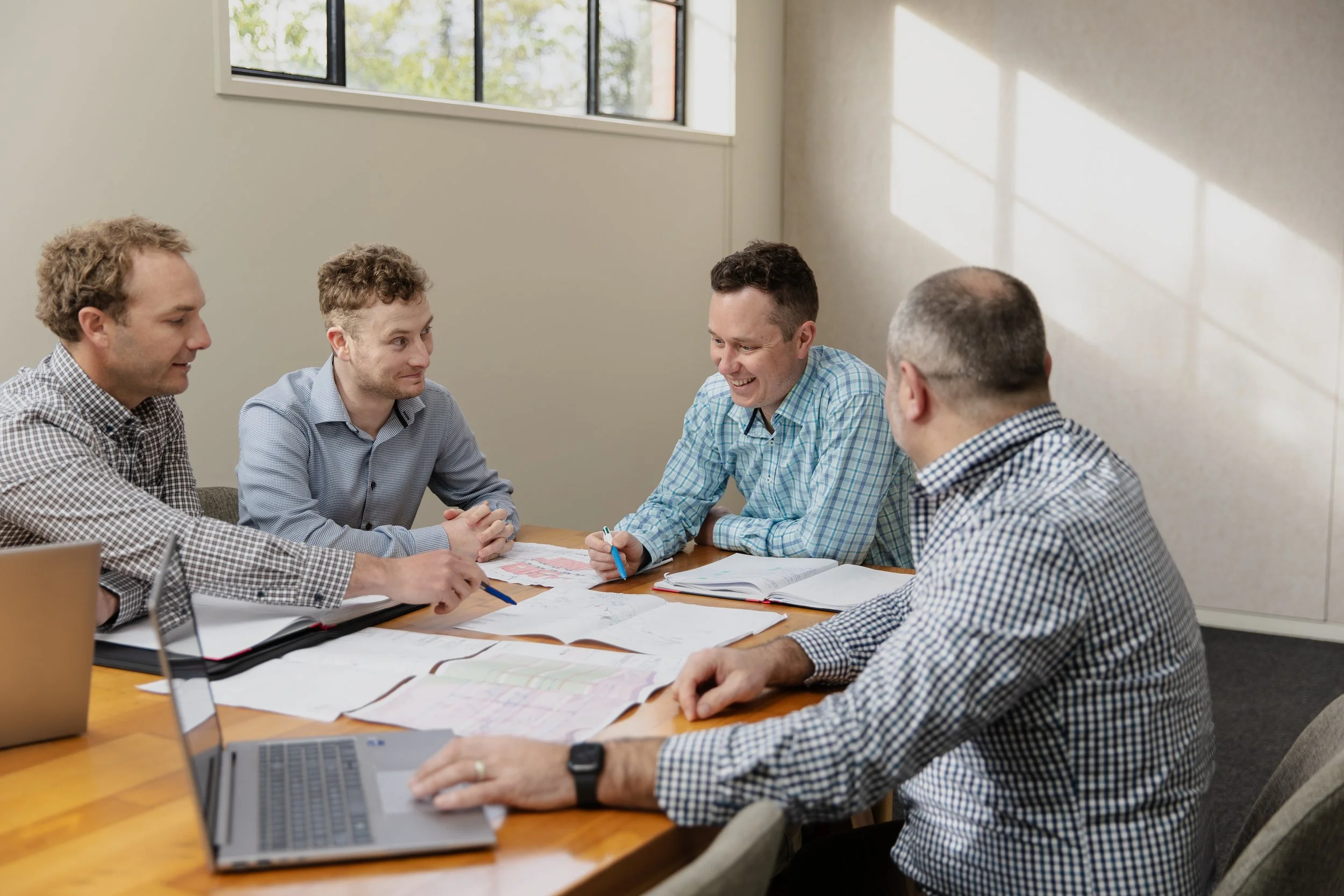 A group of five men are sitting around a wooden conference table with papers, notebooks, and laptops, engaged in a discussion in a room with a large window letting in natural light.
