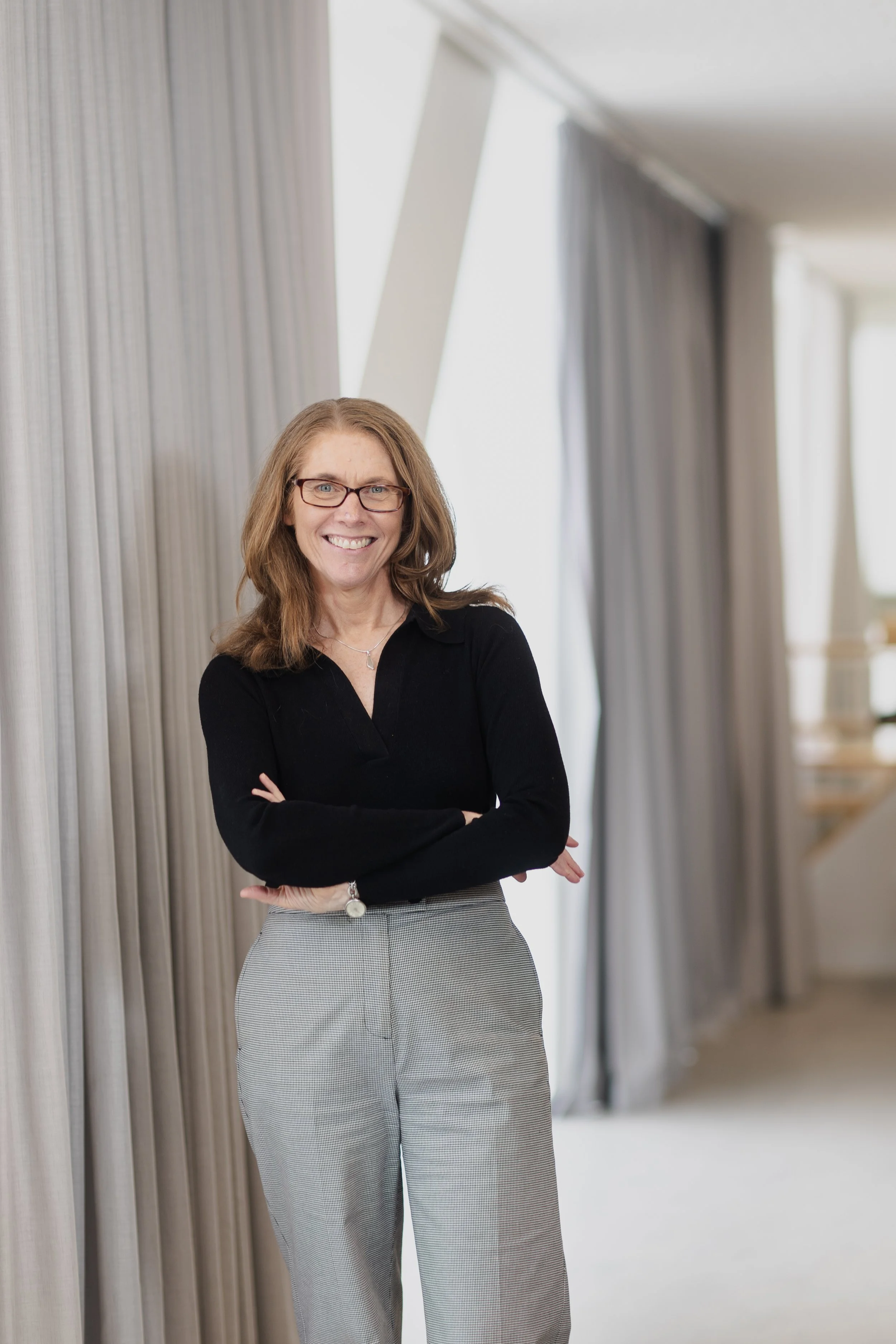 A woman with glasses, smiling and crossing her arms, standing in a bright, modern room with curtains and large windows.