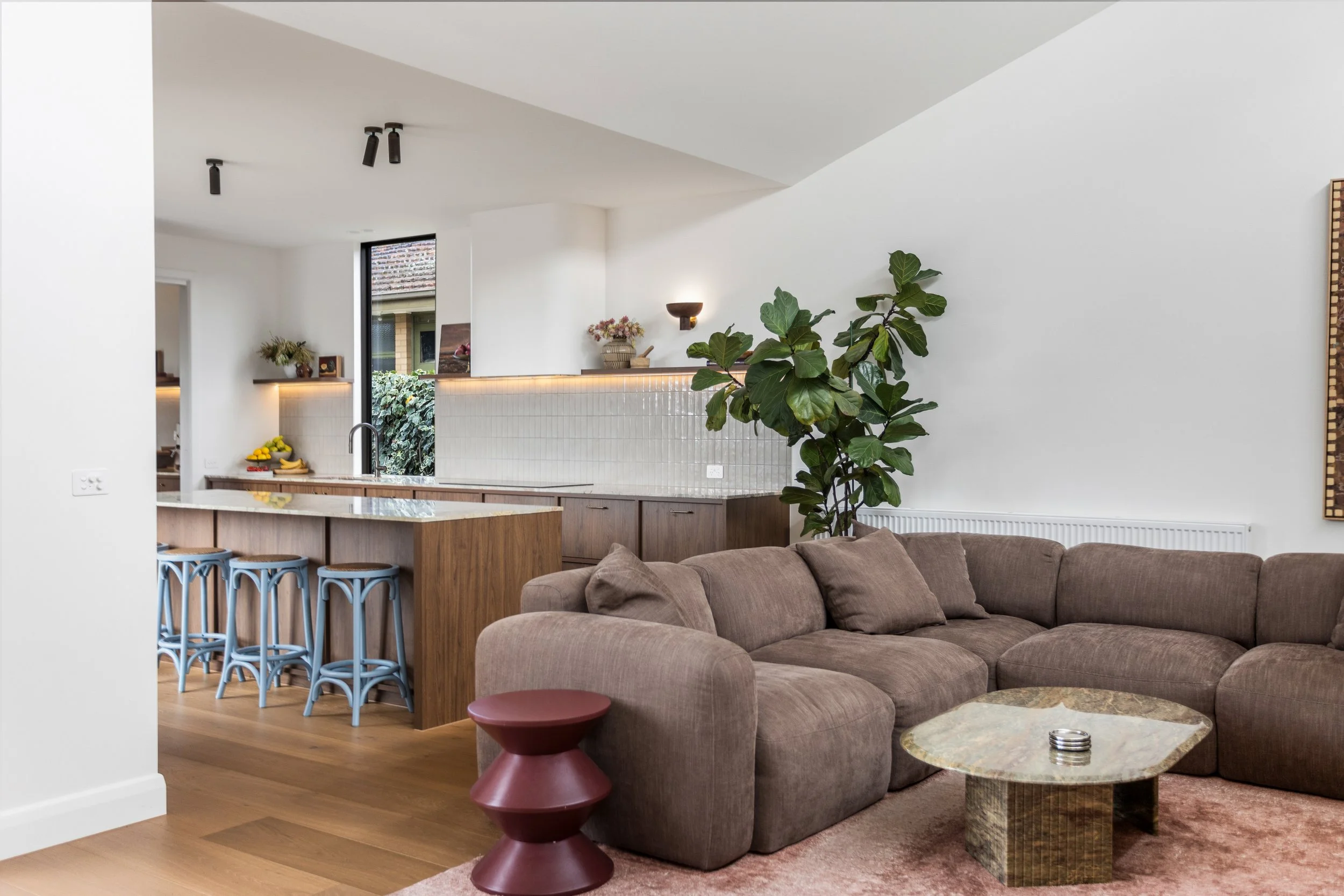 Living room with a large brown sectional sofa, a unique layered red side table, a marble coffee table, a large green plant, and a kitchen area with a wooden island, blue bar stools, and decor including flowers and baskets. The room has white walls, w