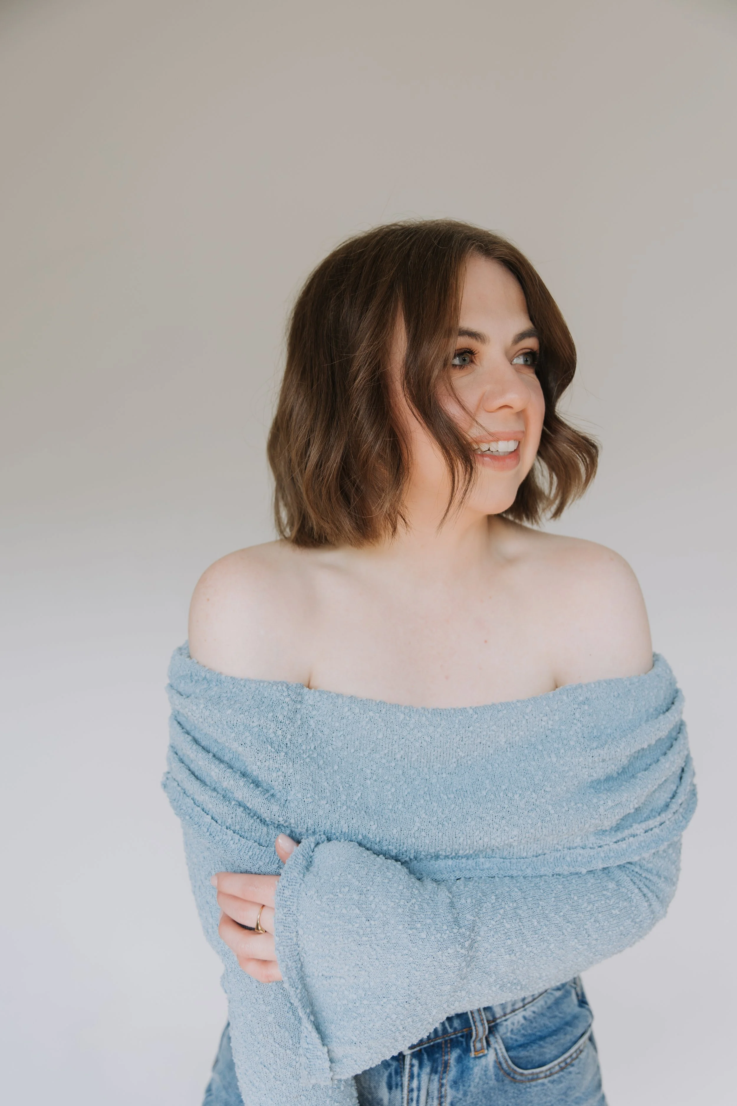 Portrait of a woman with shoulder-length wavy brown hair, smiling, wearing a light blue off-the-shoulder sweater and jeans, standing against a plain light-colored background.
