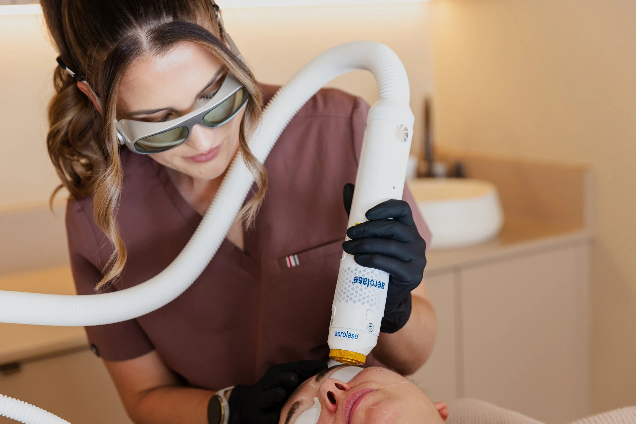 A woman wearing sunglasses and black gloves performs a cosmetic procedure on a person's face using a device labeled 'aerolase'. The person receiving treatment is lying down with eyes closed, and the setting appears to be a clinic or spa with neutral-
