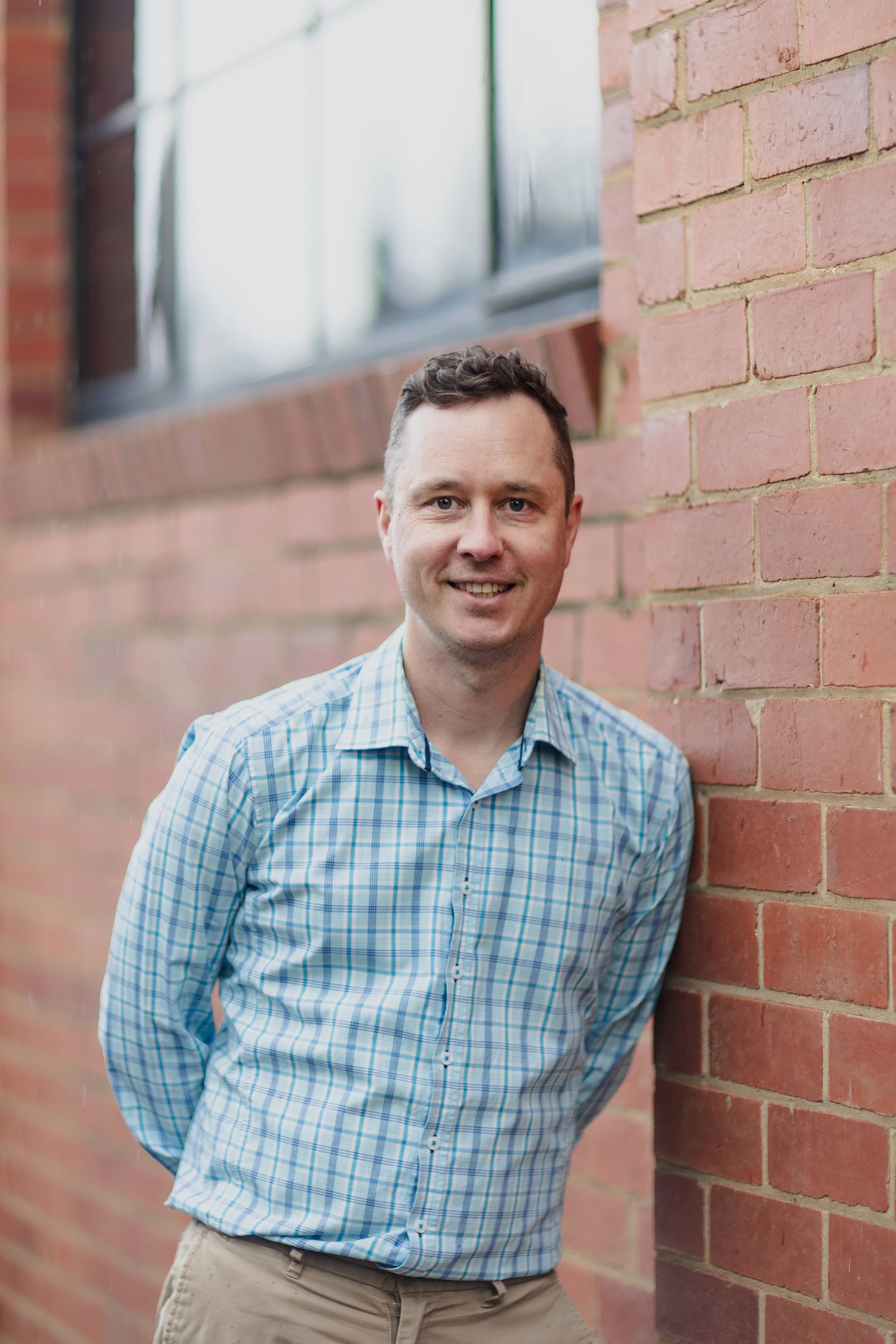 A man leaning against a brick wall next to a window, smiling, wearing a blue plaid shirt and khaki pants.