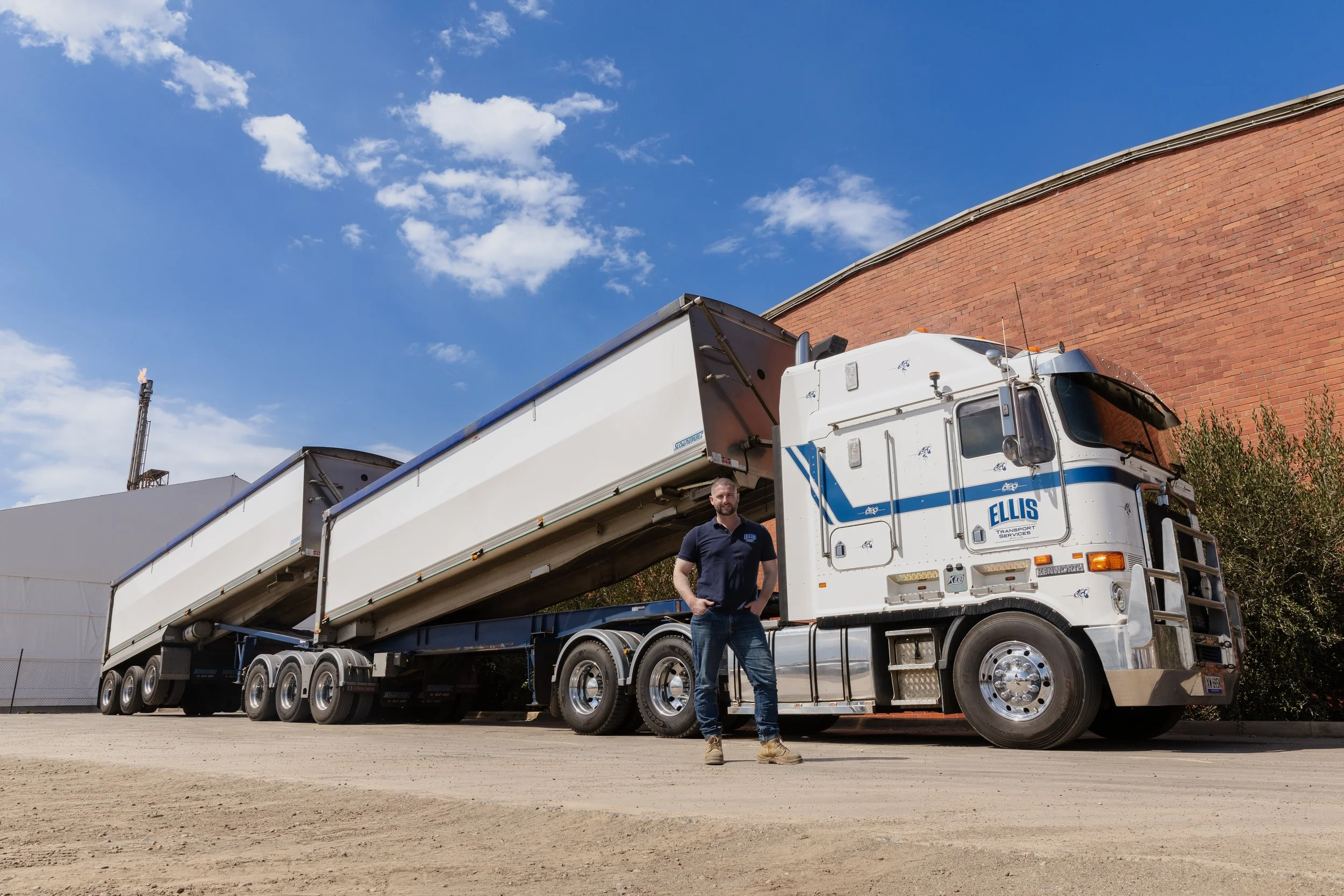 A man standing in front of a large white and blue semi-truck with an extended trailer, out in an industrial area with a brick wall and blue sky with clouds.