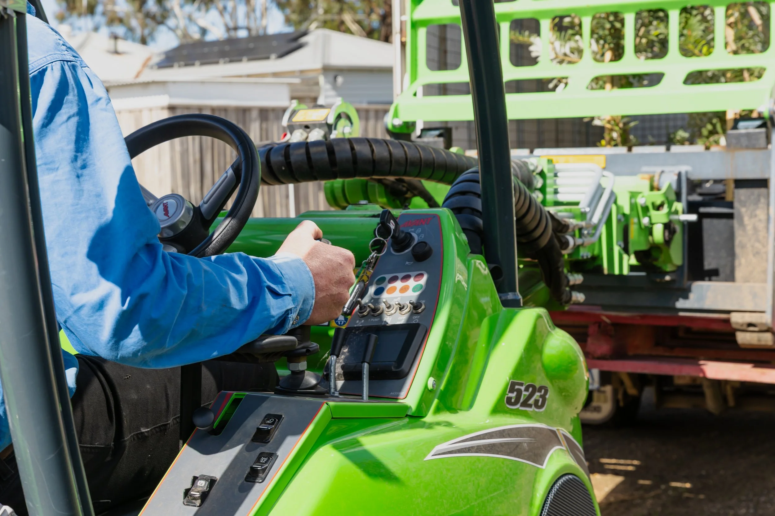 Close-up of a person driving a bright green utility vehicle with control panel and black hoses, outdoors with a wooden fence and buildings in the background.
