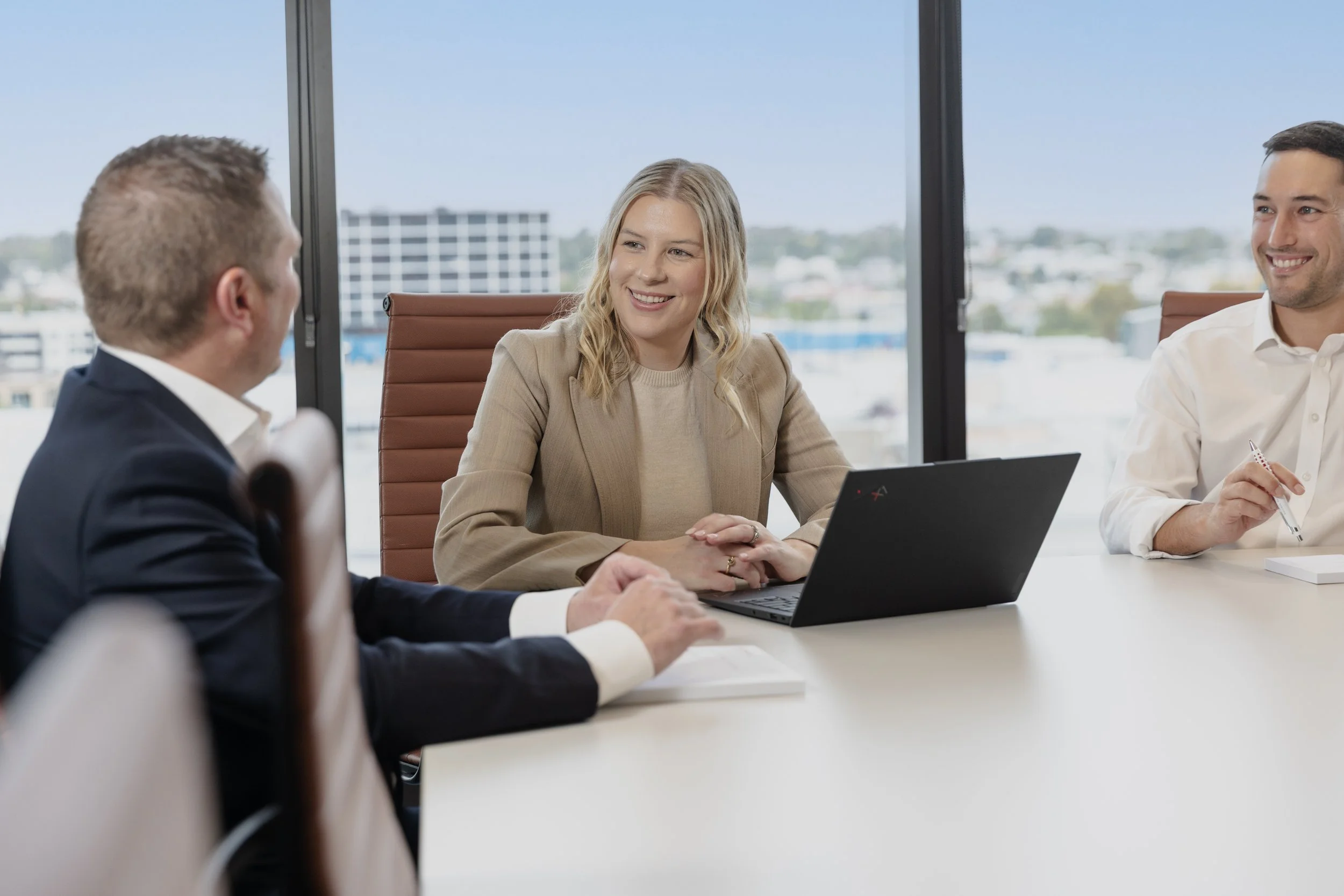 Three business professionals sitting around a conference table, engaging in a discussion, with a cityscape visible through the large windows behind them.