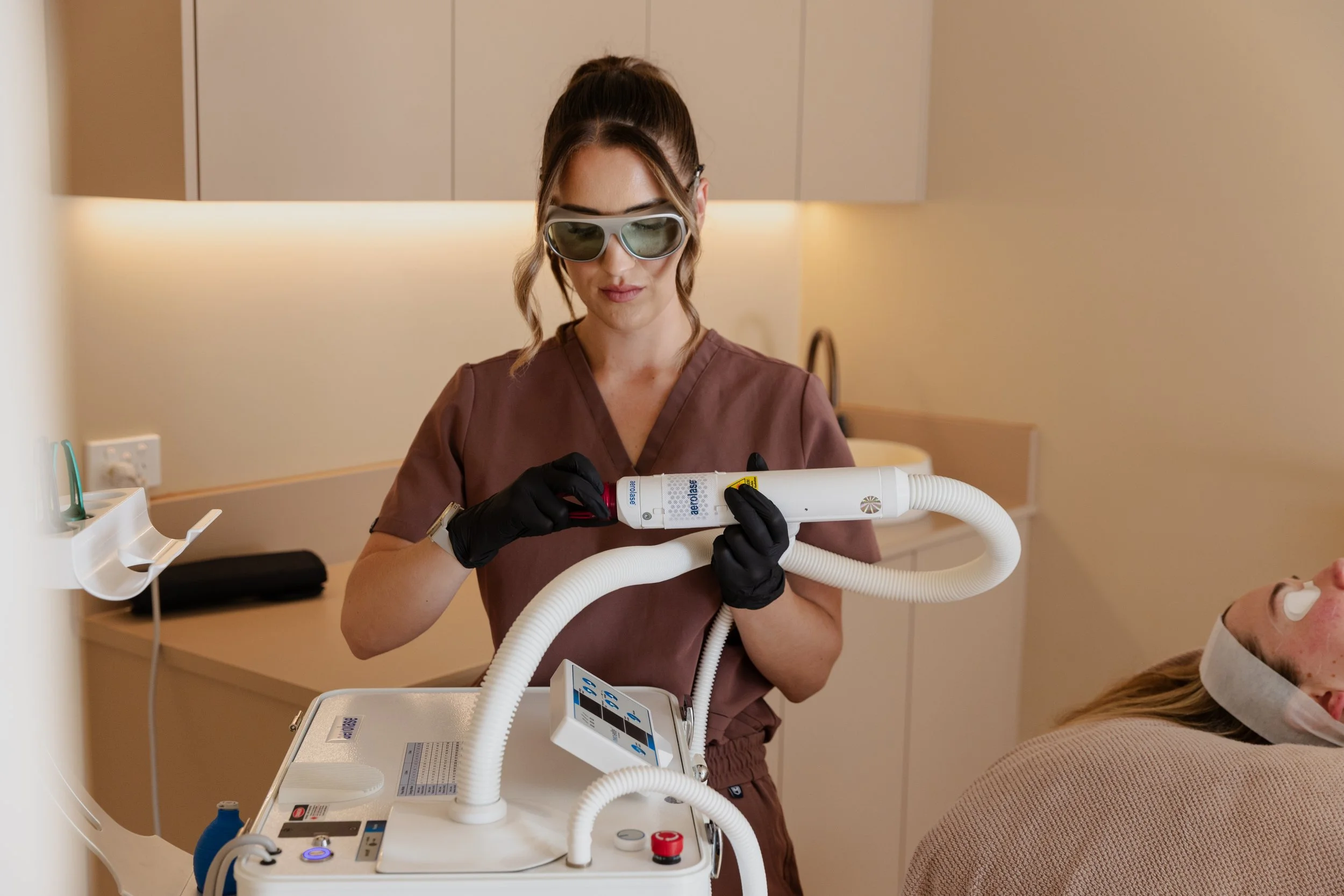 A healthcare professional in protective glasses and black gloves operating a medical device on a patient lying on a chair in a clinical setting.