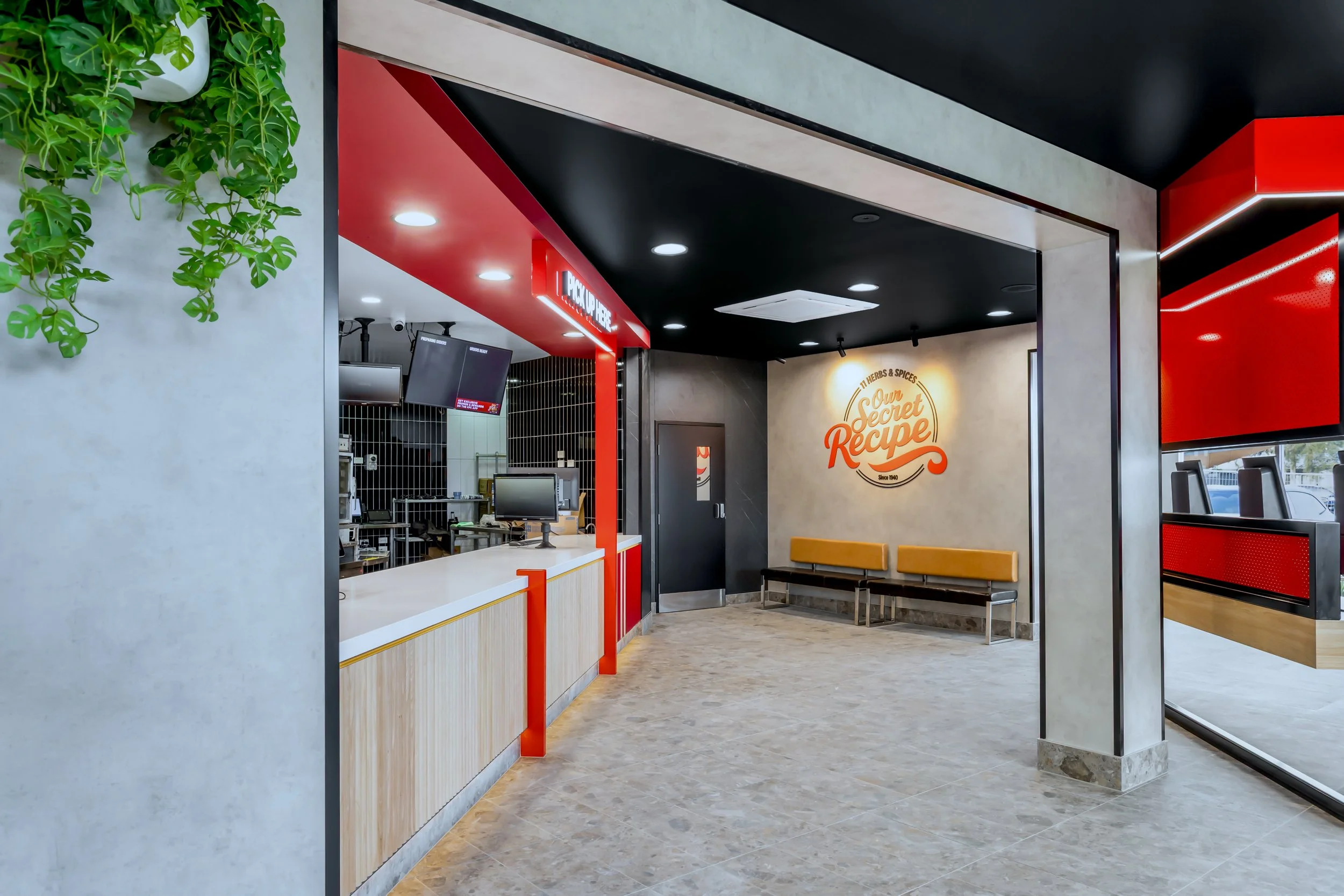 Modern fast-food restaurant interior with a red and black color scheme. There is a counter with digital menu screens, a seating area with wooden benches, and a wall with the restaurant's logo "Our Secret Recipe" in orange and white. Green plants are 