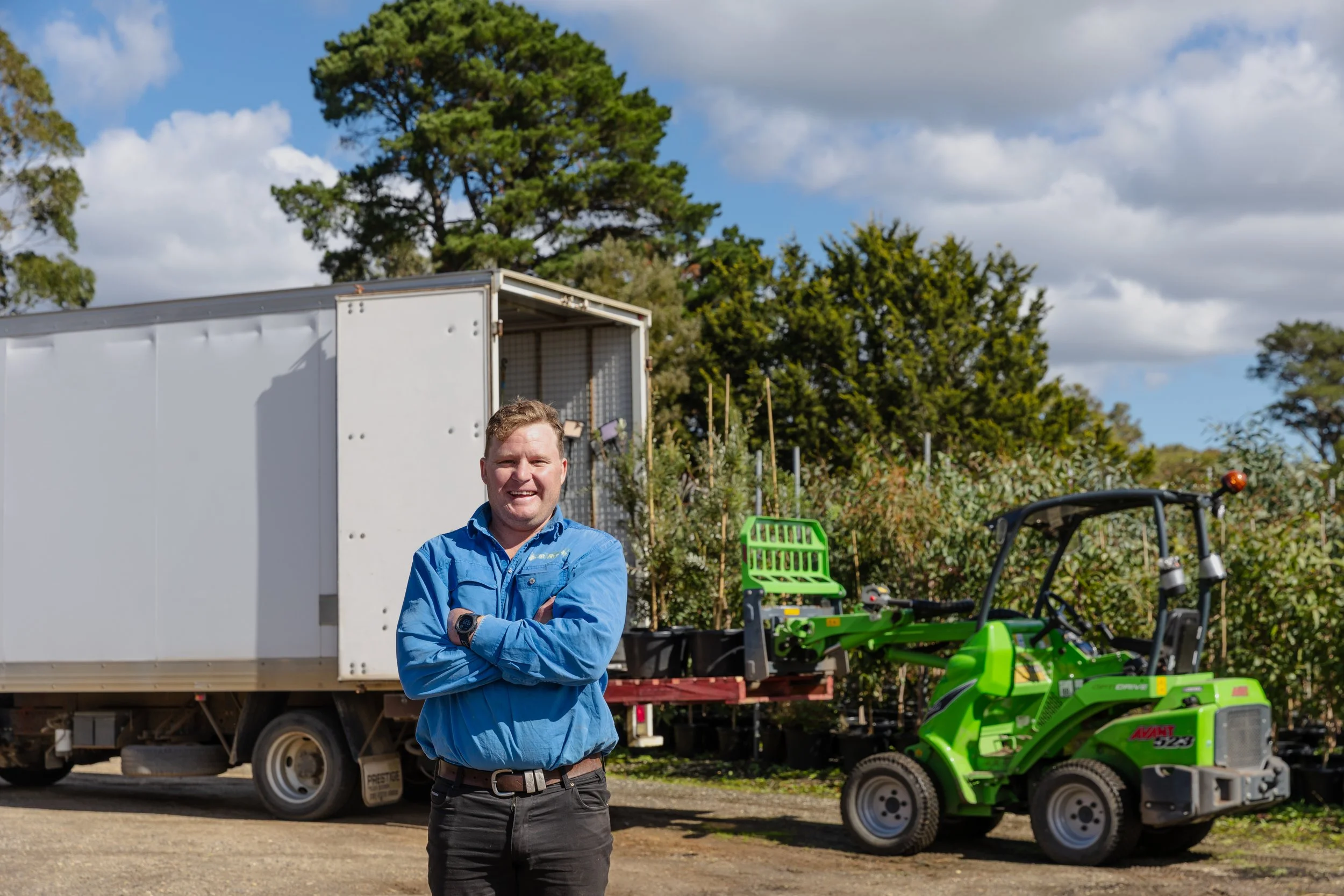 A man in a blue shirt and black pants standing outdoors with arms crossed, smiling. Behind him are trees, a large white truck, and green landscaping equipment, under a partly cloudy sky.