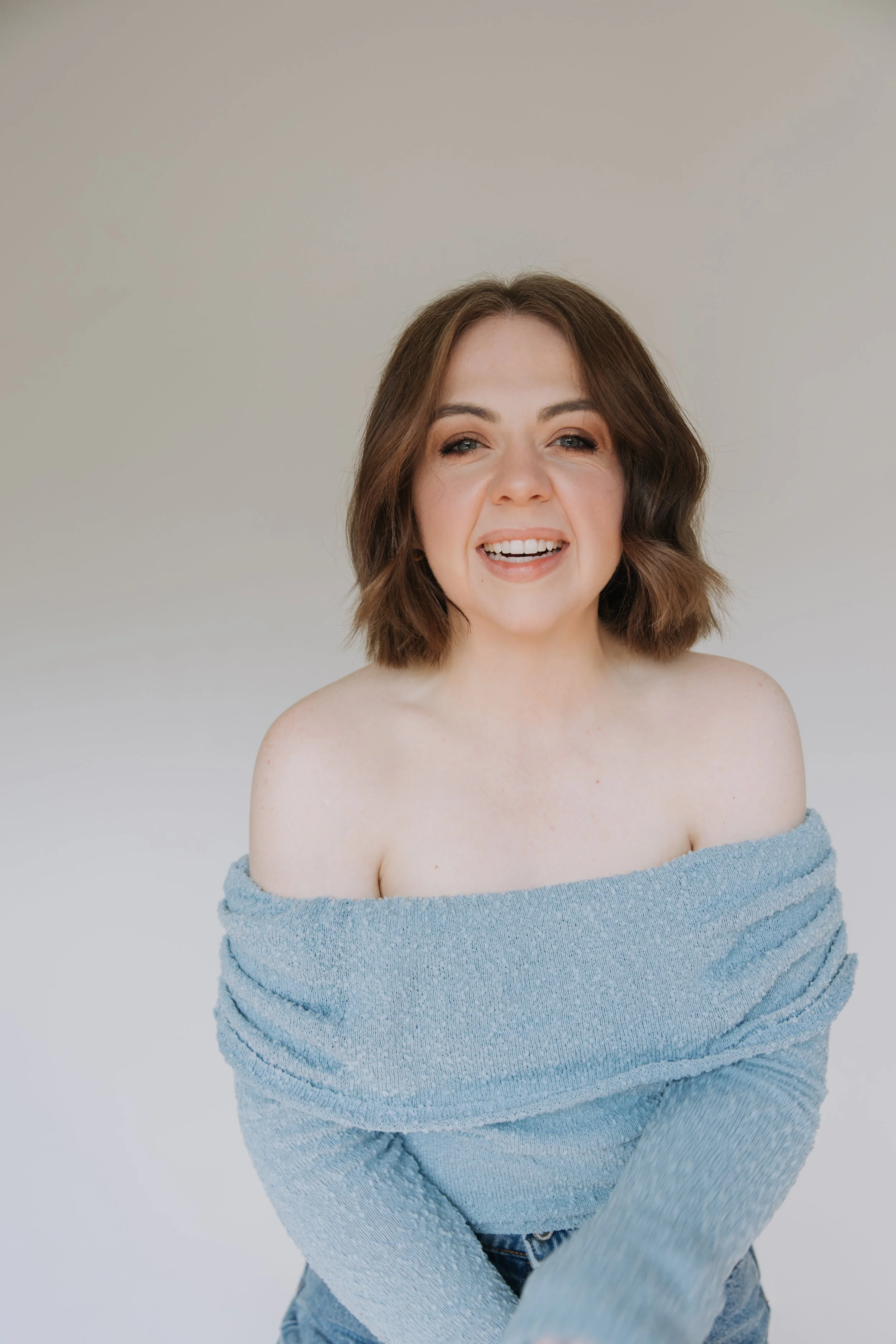 A woman with shoulder-length brown hair smiling, wearing a light blue off-the-shoulder sweater and jeans, against a plain white background.