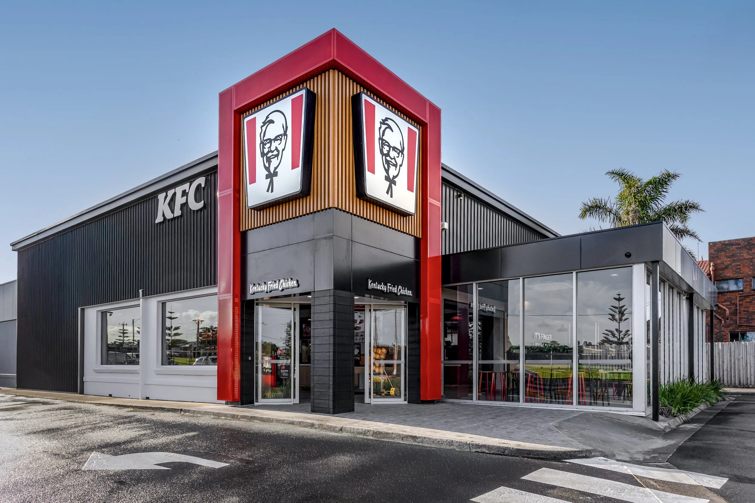 Exterior view of a KFC restaurant with a prominent Colonel Sanders logo sign on a red and black building, glass windows, and a parking lot in front, under a clear blue sky.