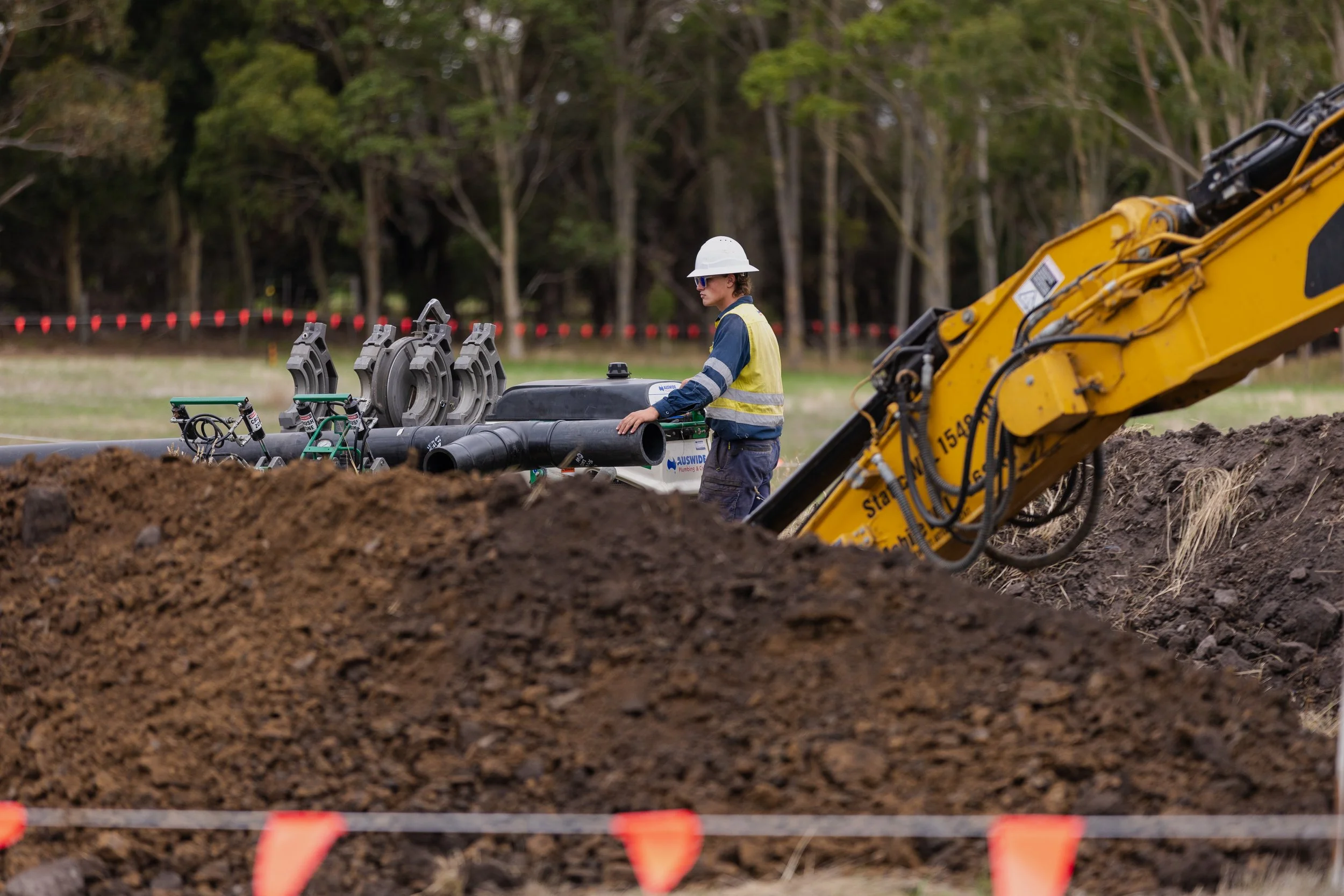 A construction worker wearing a white safety helmet and yellow reflective vest operating heavy machinery at a construction site surrounded by dirt and trees in the background.