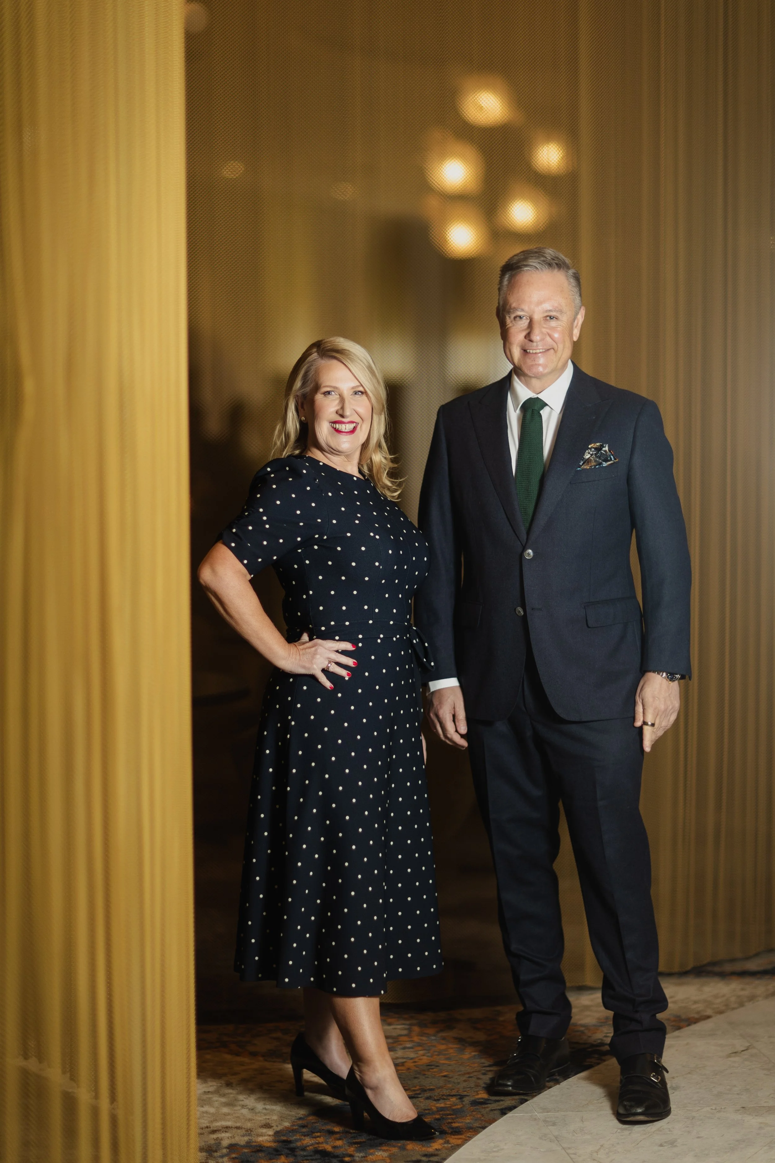 A woman and man standing together indoors, smiling for the camera. The woman is wearing a navy blue polka dot dress and black high heels, while the man is dressed in a dark suit, white shirt, and green tie. The background features warm lighting and v