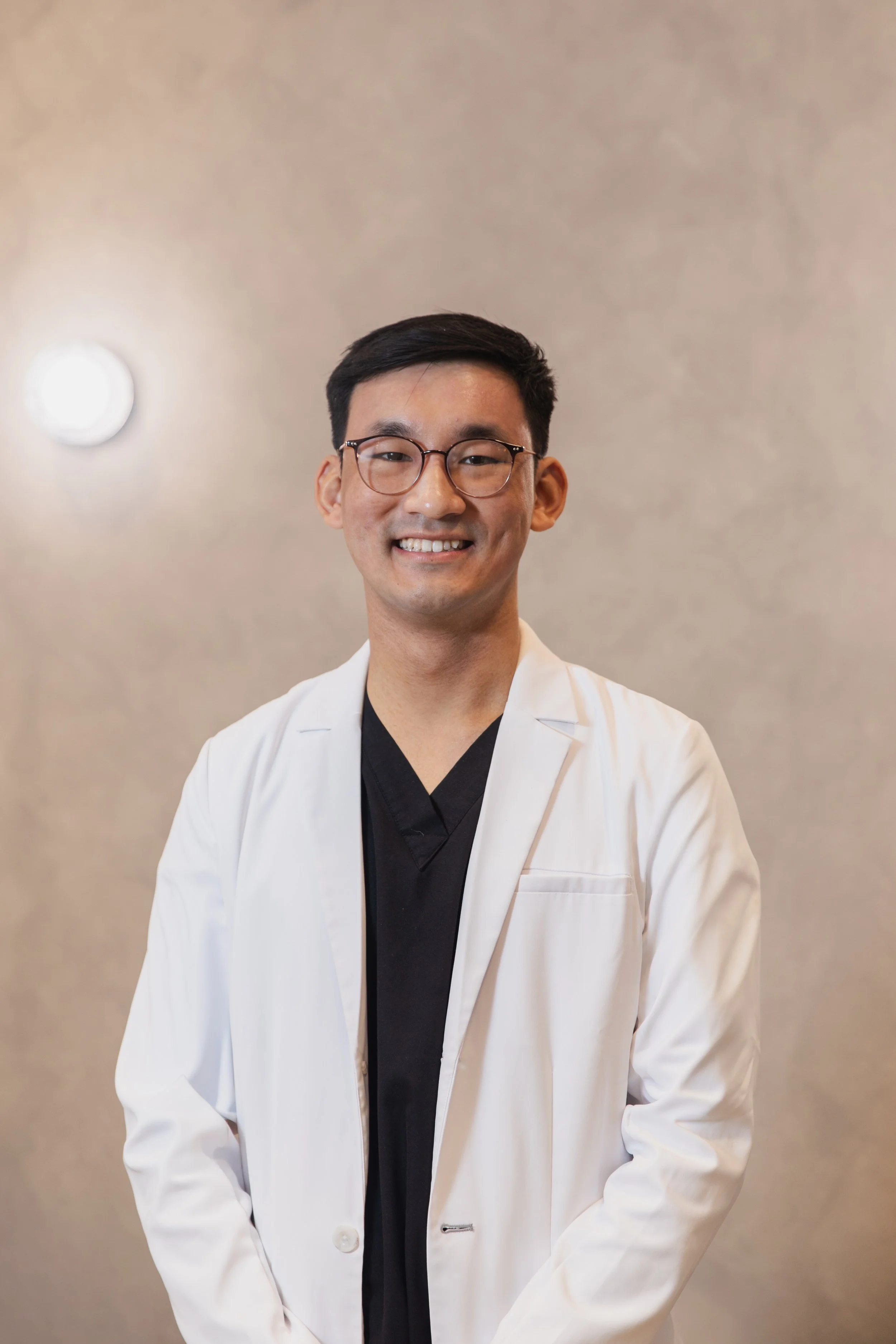 A young man with black hair, glasses, and a white medical coat smiling.