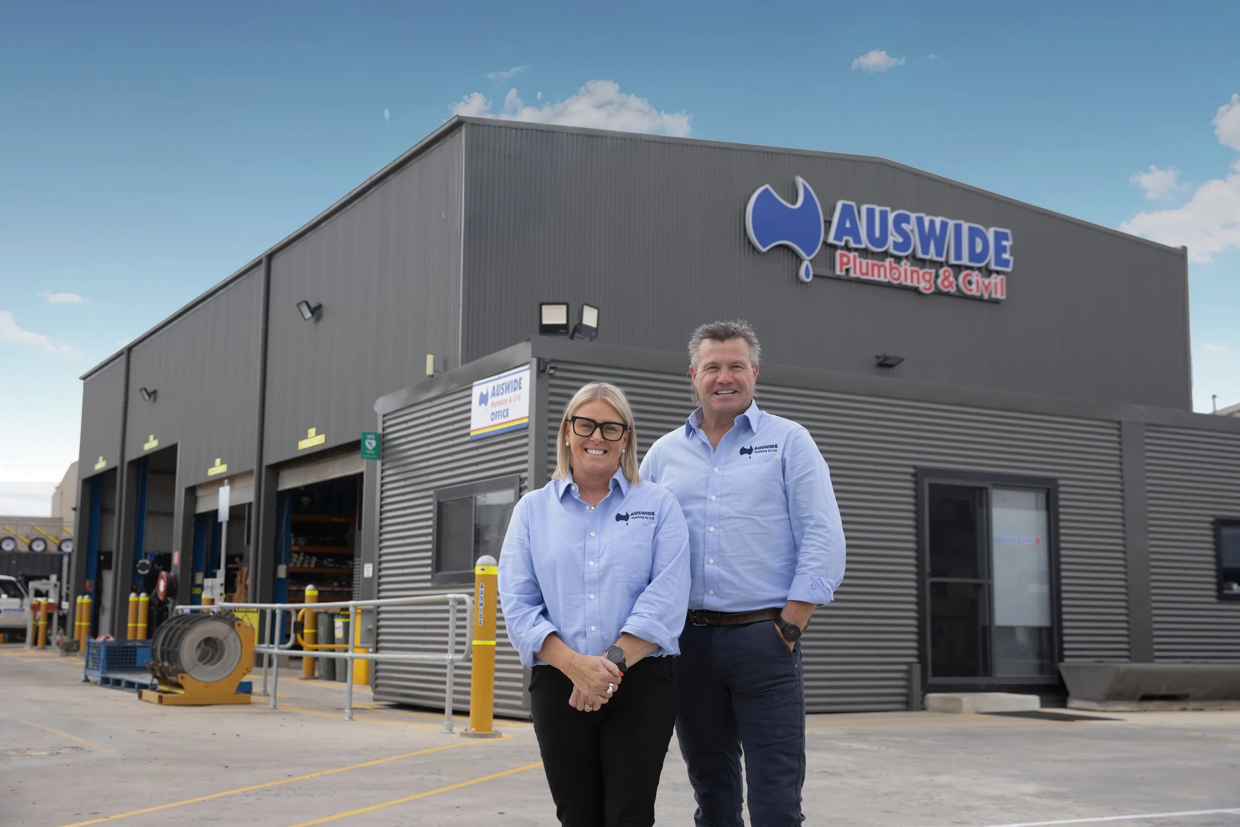 Two professionals standing outside an AUSWIDE Plumbing & Civil business building, smiling at the camera, in work uniforms with company logos, in a parking lot with tools and equipment visible.