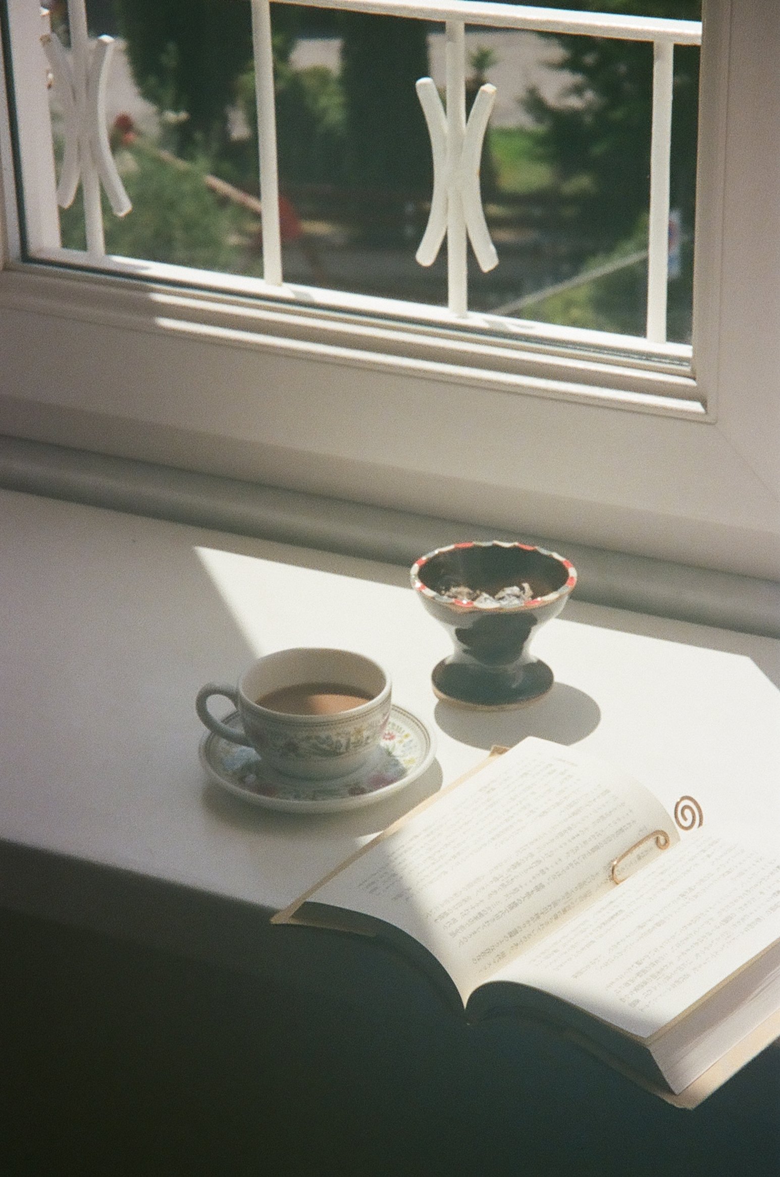 A windowsill with a cup of tea or coffee in a patterned cup and saucer, an open book, and a small decorative bowl, all bathed in sunlight with a view of green trees outside.