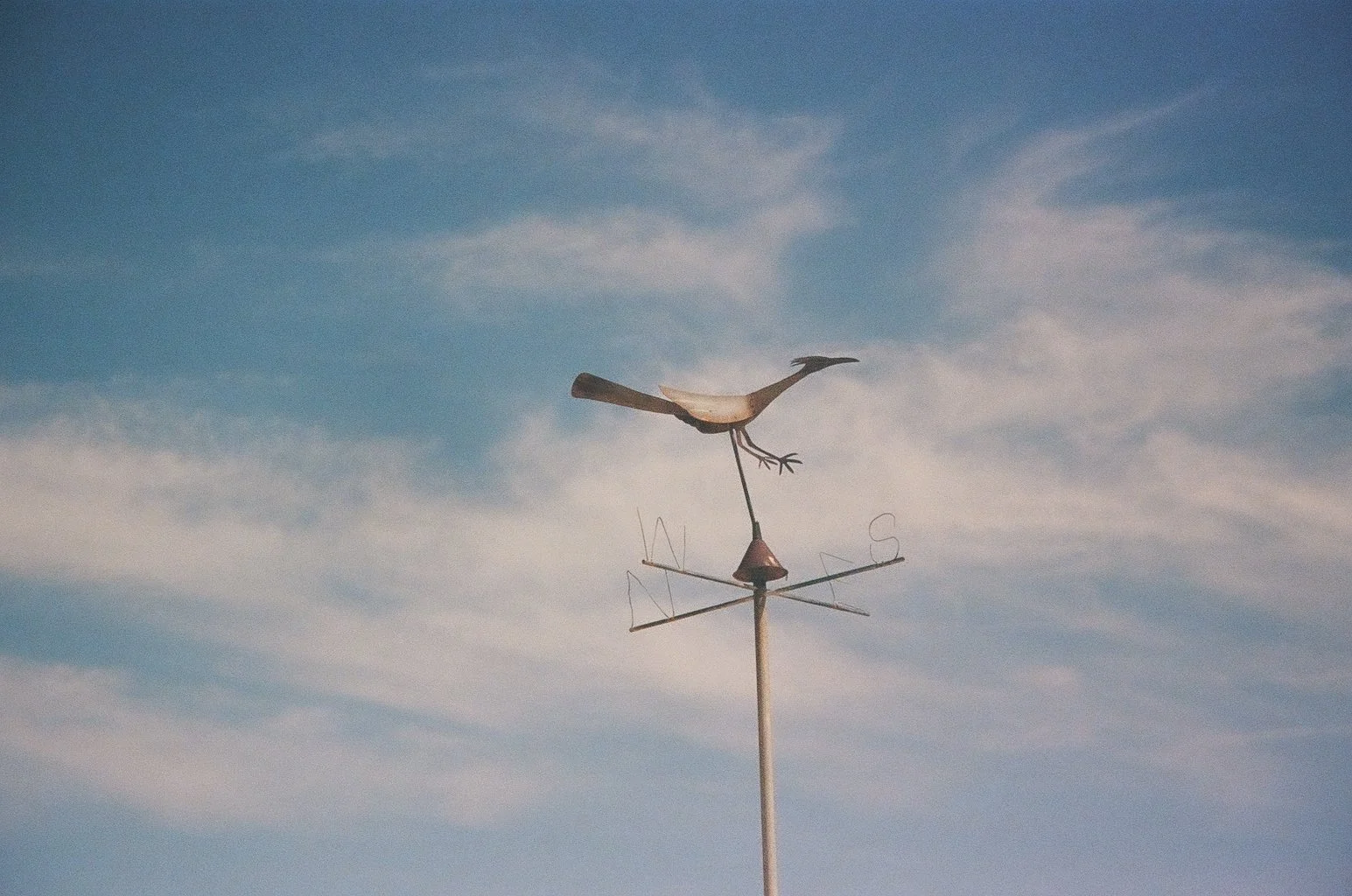 A metal weather vane with a bird figure on top, showing the directions N, E, S, W against a blue sky with wispy clouds.
