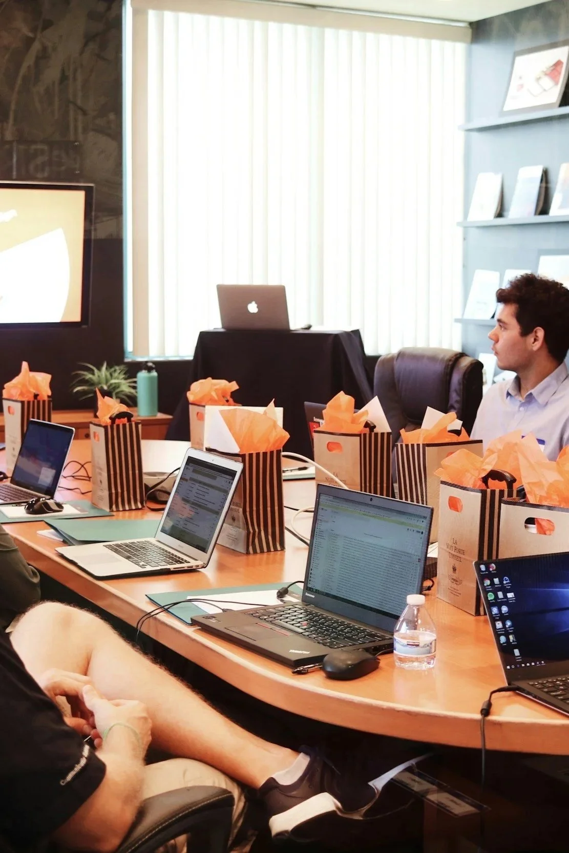 Business meeting room with laptops on a conference table, gift bags with orange tissue paper, a water bottle, and a man sitting at the table.