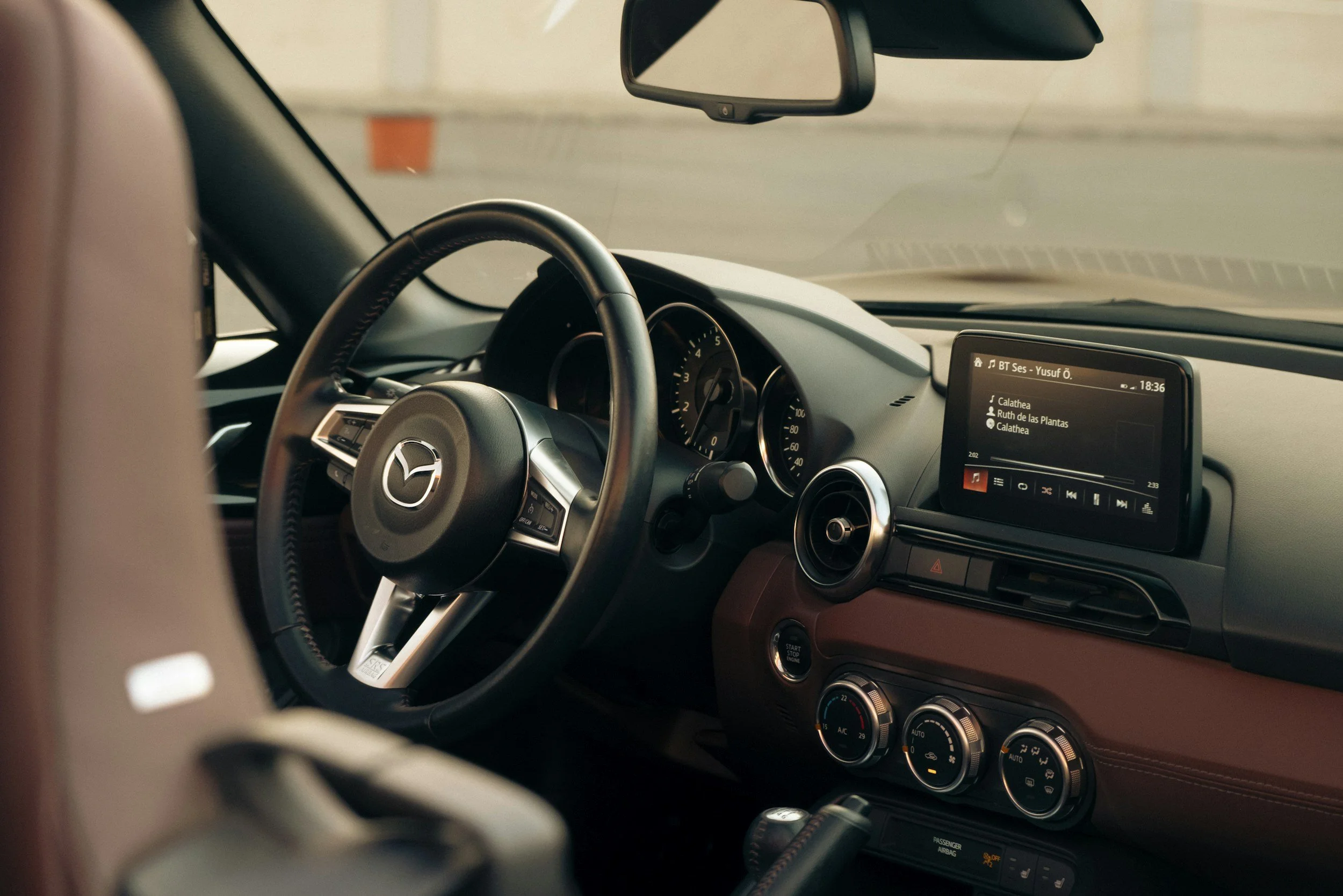 Interior view of a Mazda car dashboard, showing the steering wheel, speedometer, and infotainment screen displaying a music playlist.