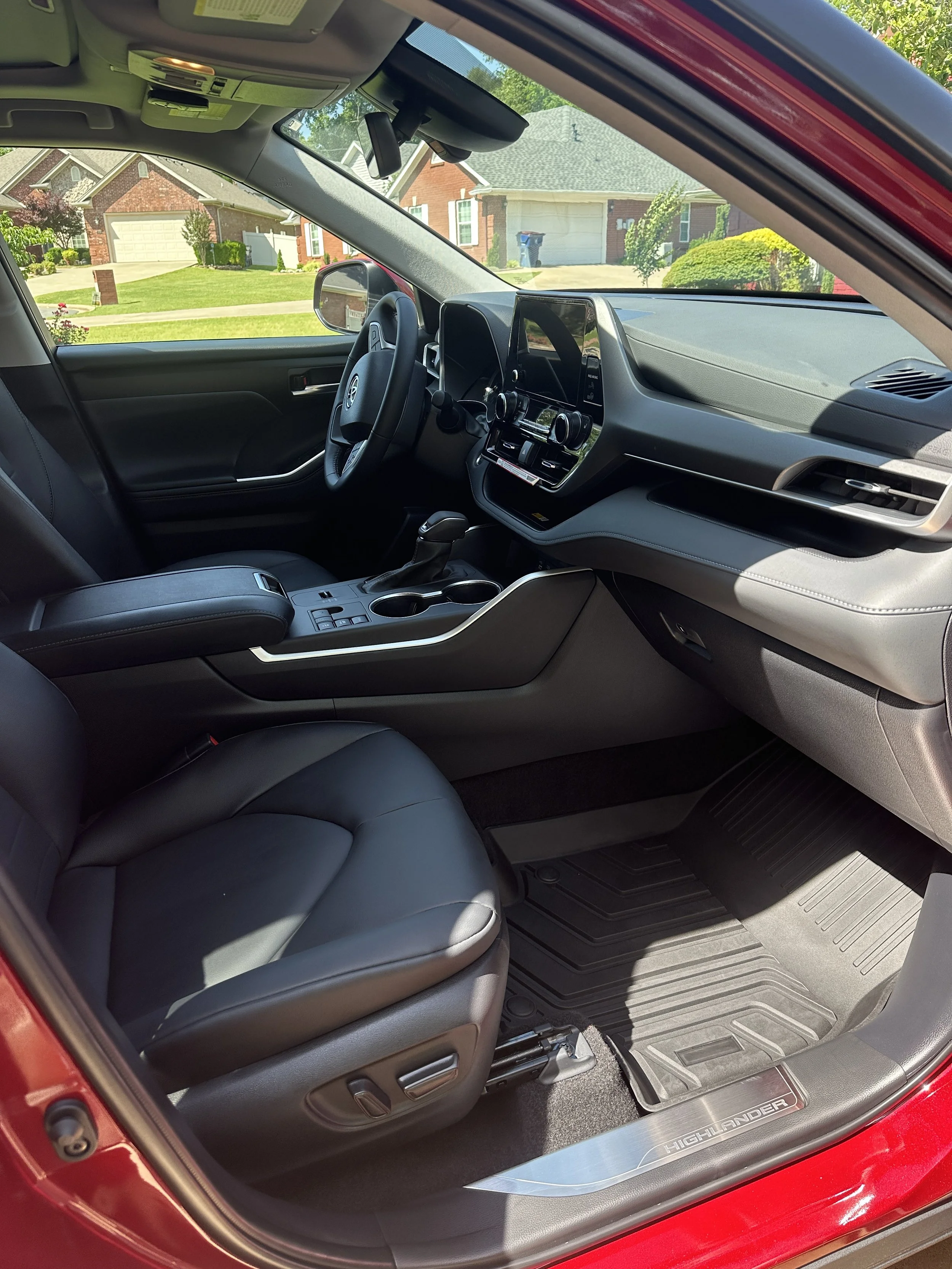 Interior of a red Toyota Highlander SUV showing the driver's seat, dashboard, steering wheel, and center console with a view of a suburban neighborhood through the windows.